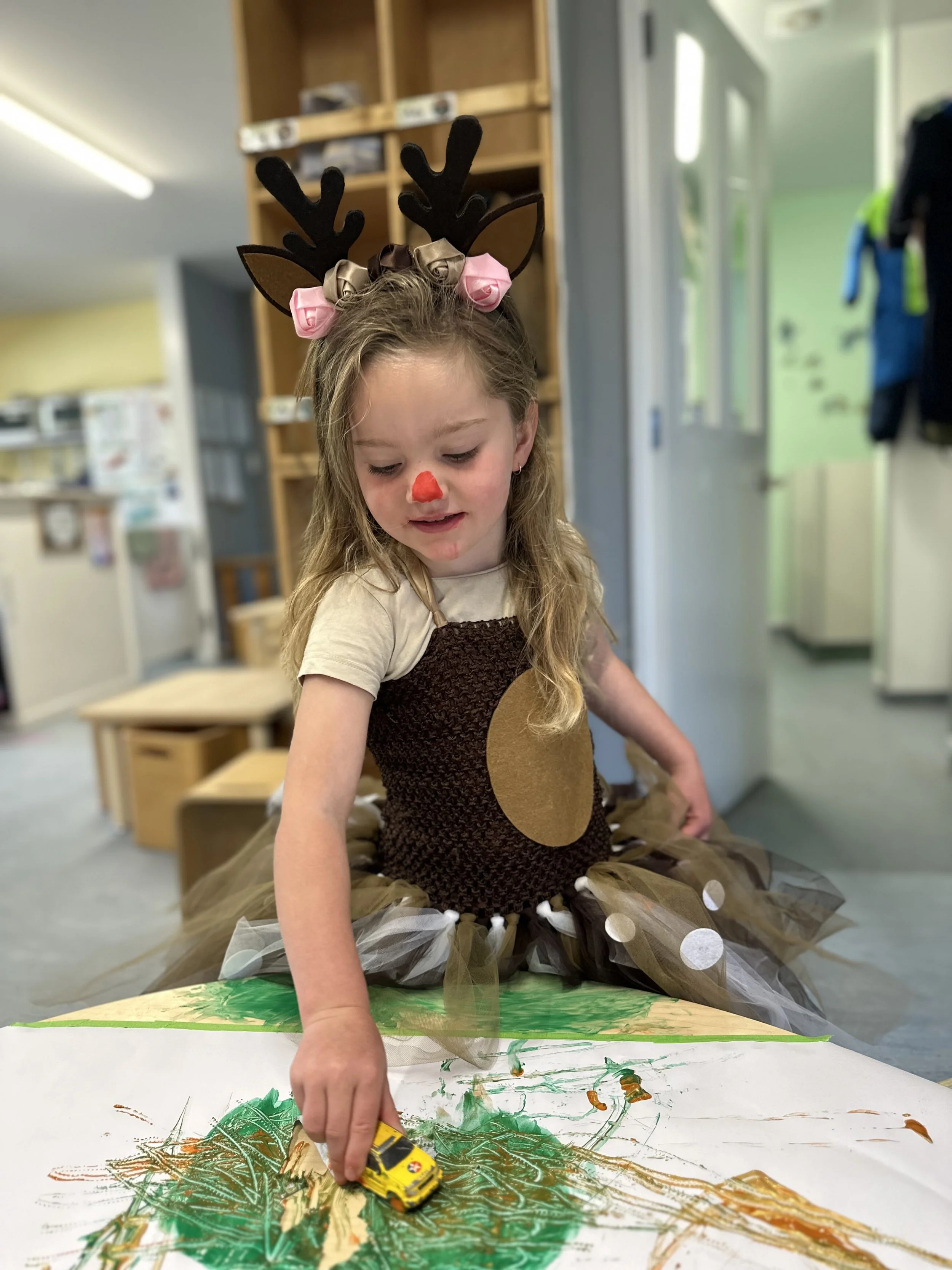 Girl dressed as a reindeer with antlers, a red nose, and pink roses on her head, painting a large colorful picture at a table in a classroom.