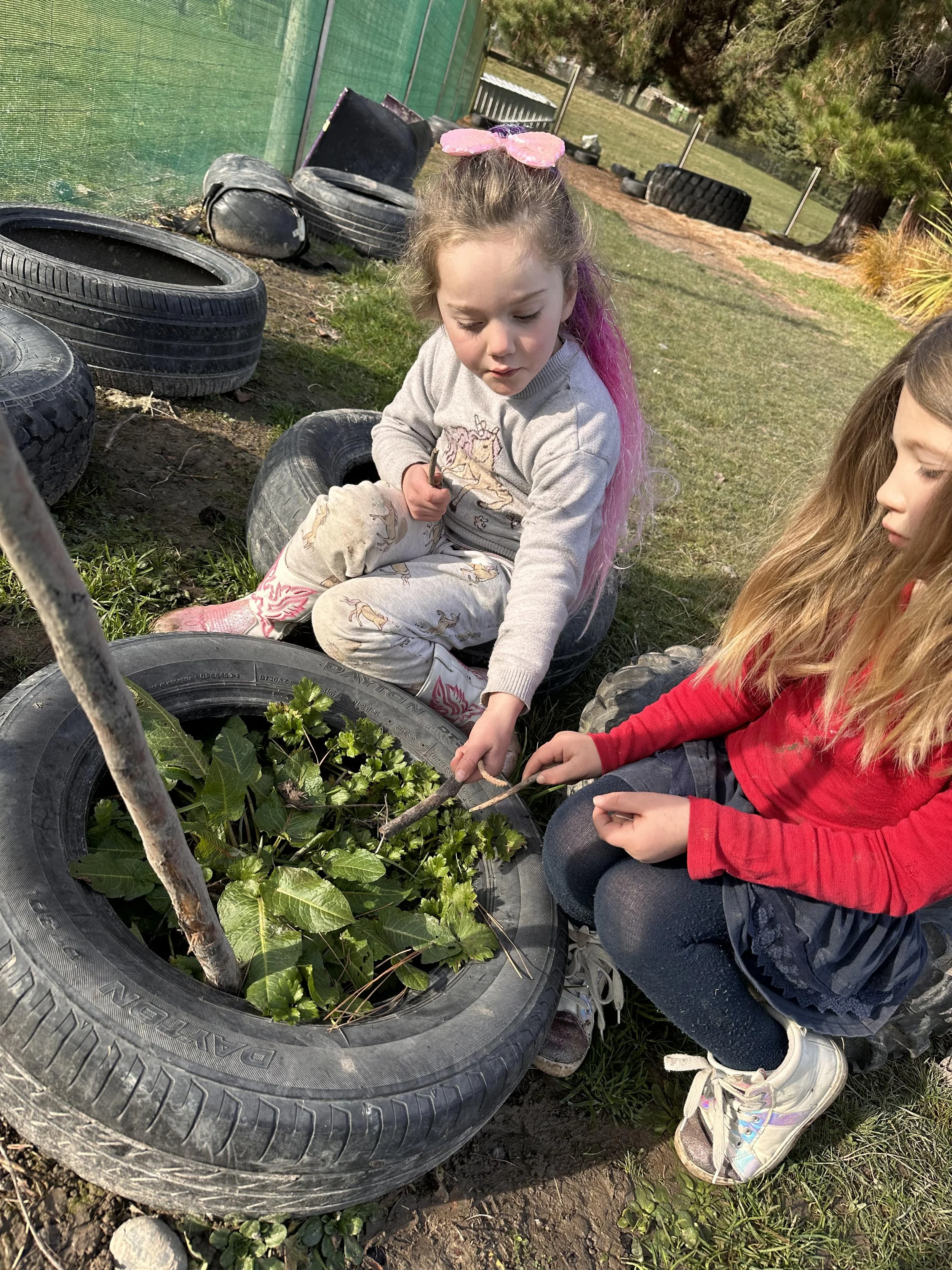 Two young girls are sitting on tires arranged as garden planters with plants growing inside, outdoors on a grassy area, surrounded by trees and tires.