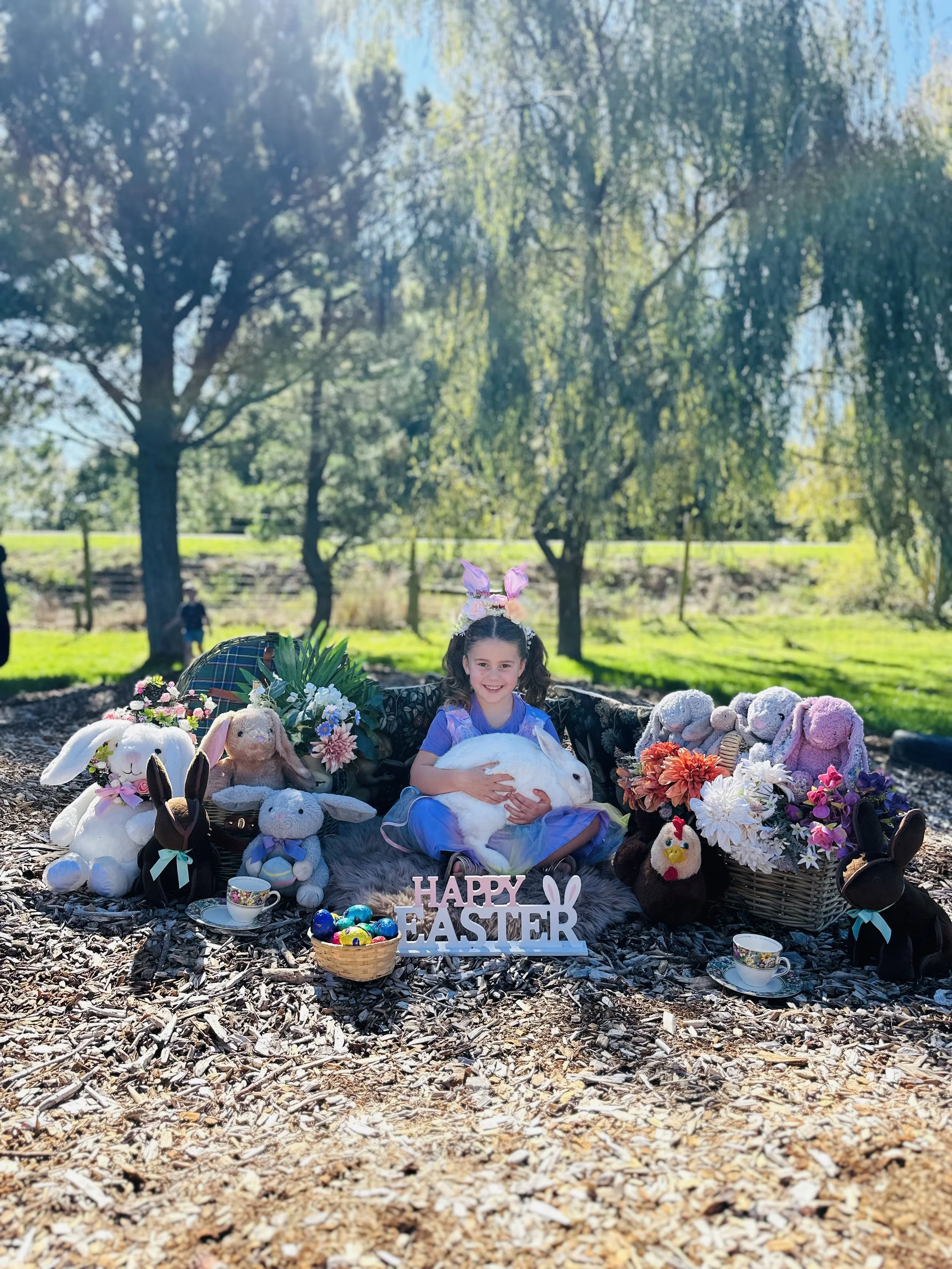 A girl sitting outdoors surrounded by stuffed animals, flowers, and Easter decorations, with a sign that says 'HAPPY EASTER' in front of her.