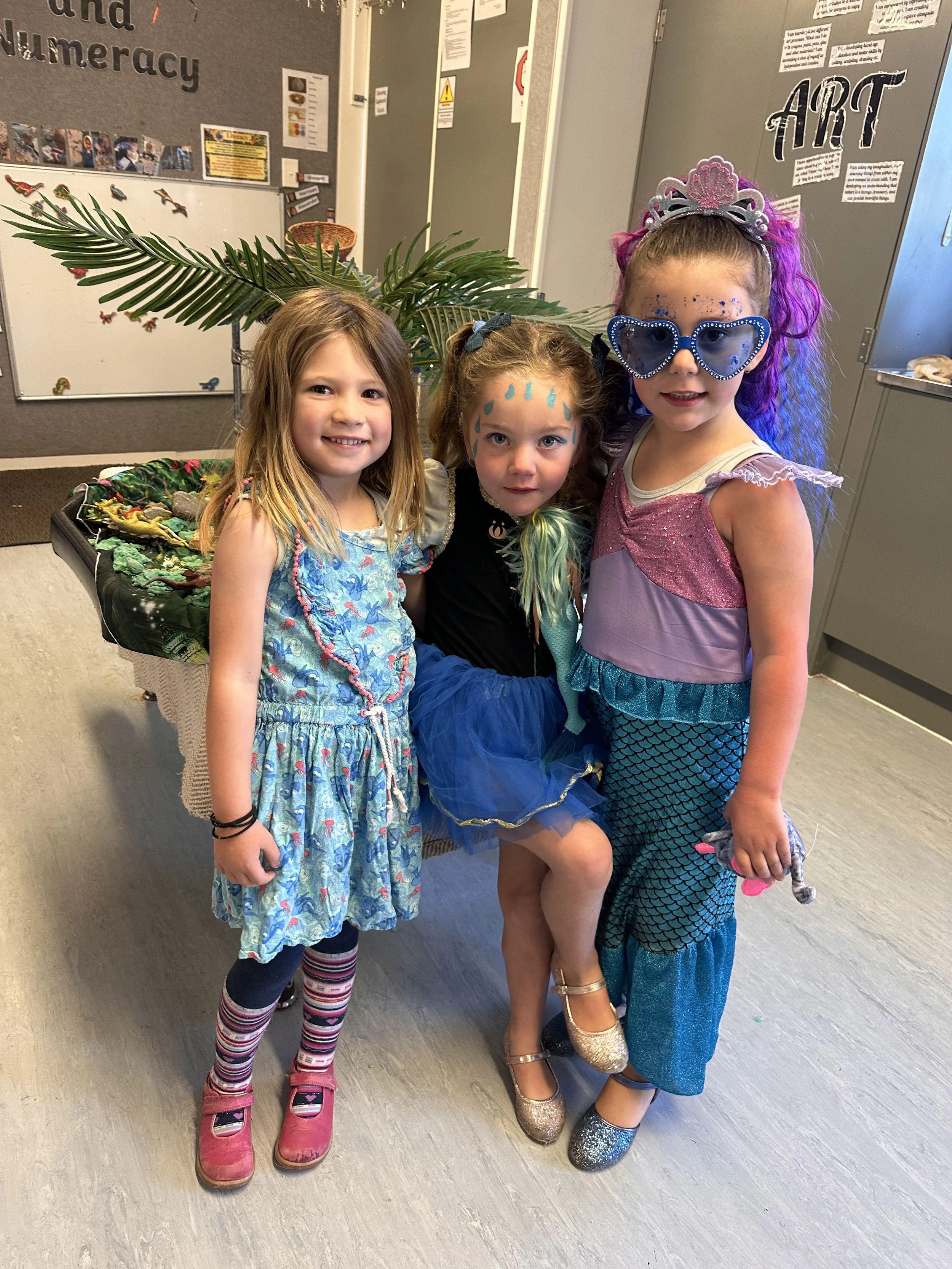 Three young girls in colorful costumes and accessories, standing indoors in front of a gray wall with art displays. One girl has face paint and is dressed as a mermaid, the second girl is wearing a blue dress with striped tights, and the third girl i