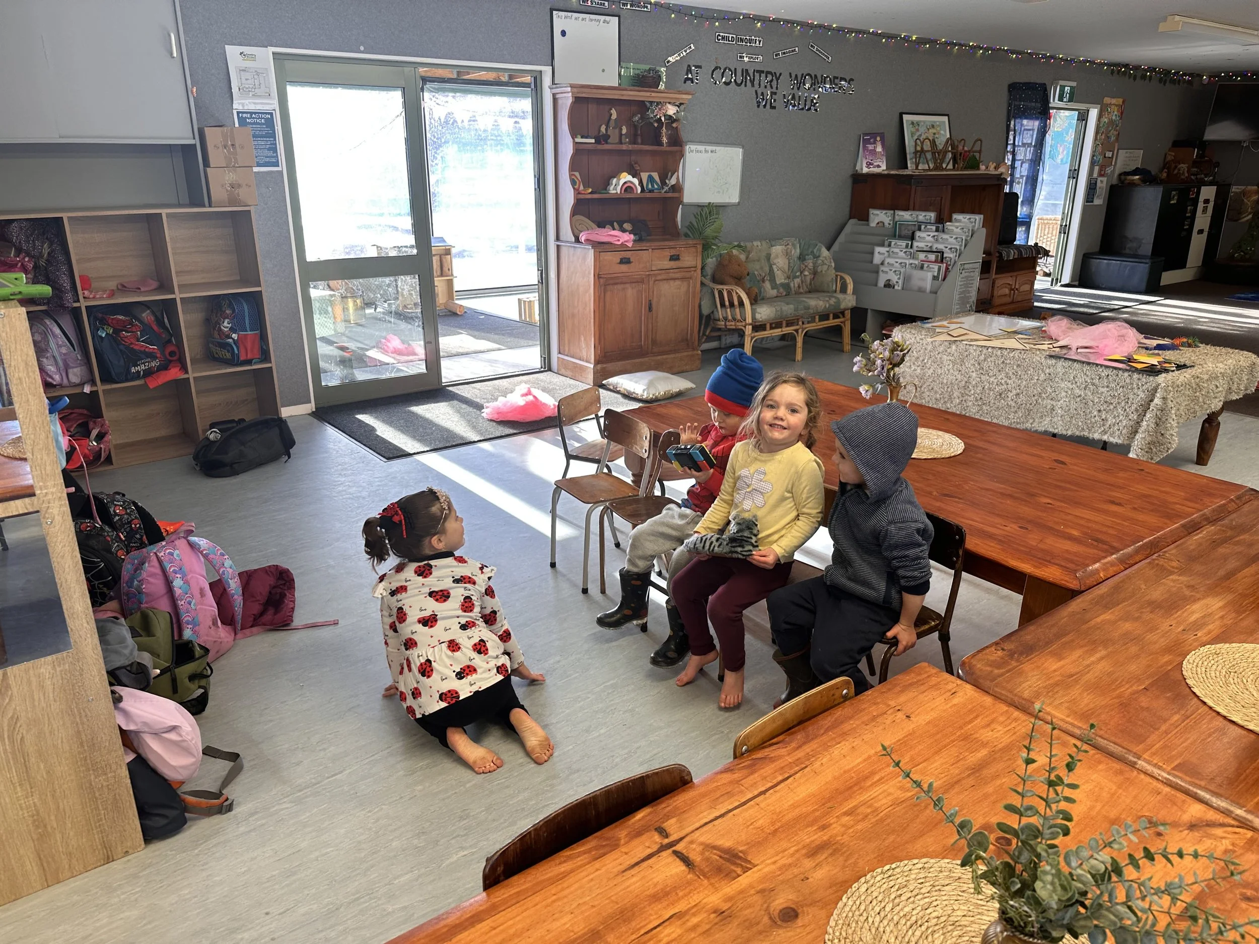 A group of children is sitting and standing around a wooden table in a brightly lit indoor space, with backpacks on the floor and smiling faces.