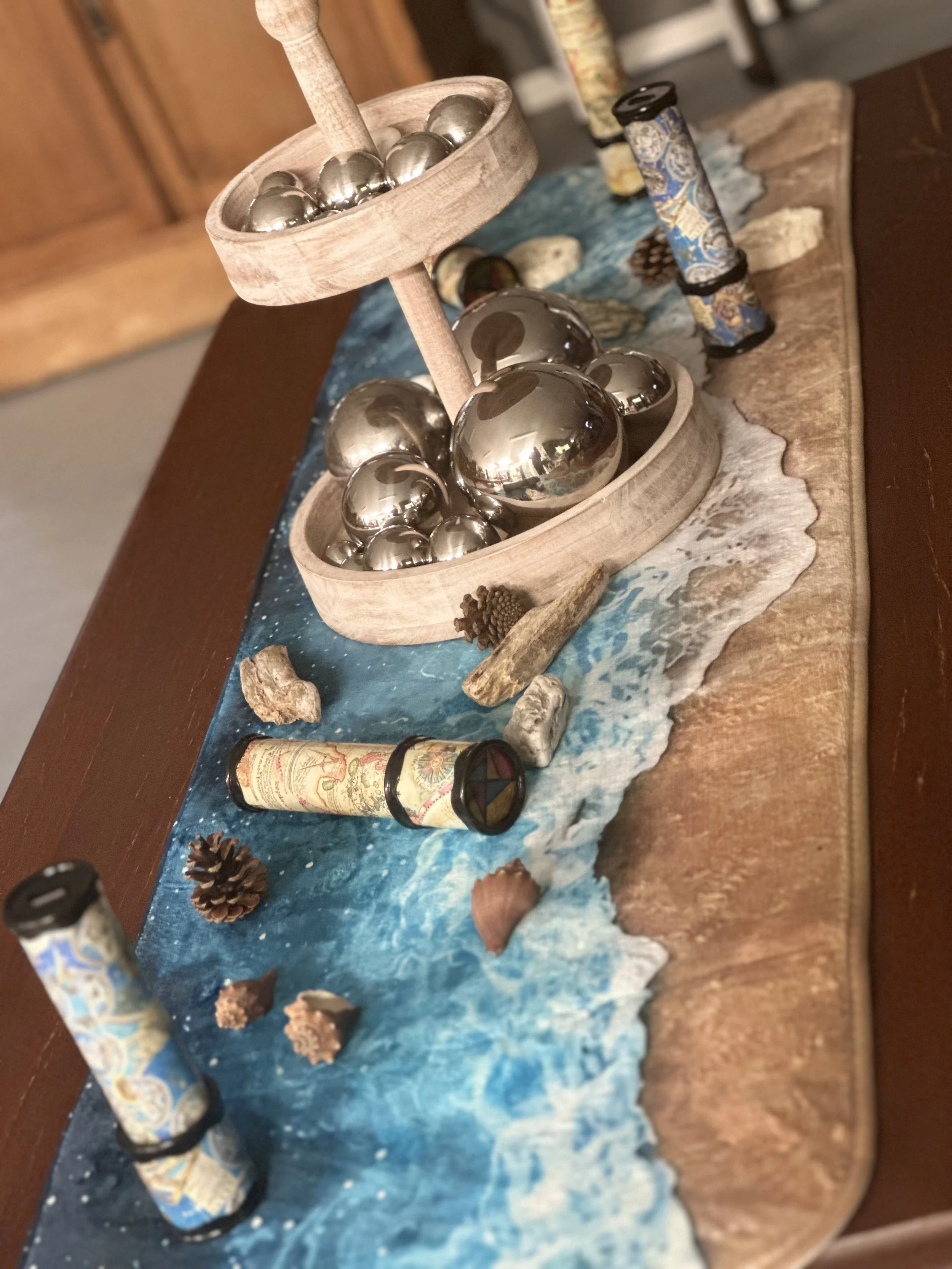 A decorative tabletop scene featuring a wooden display stand with metallic spheres and small dollops of white material, pinecones, and various decorative rolled papers, all arranged on a dark wooden surface with a piece of blue fabric or paper undern
