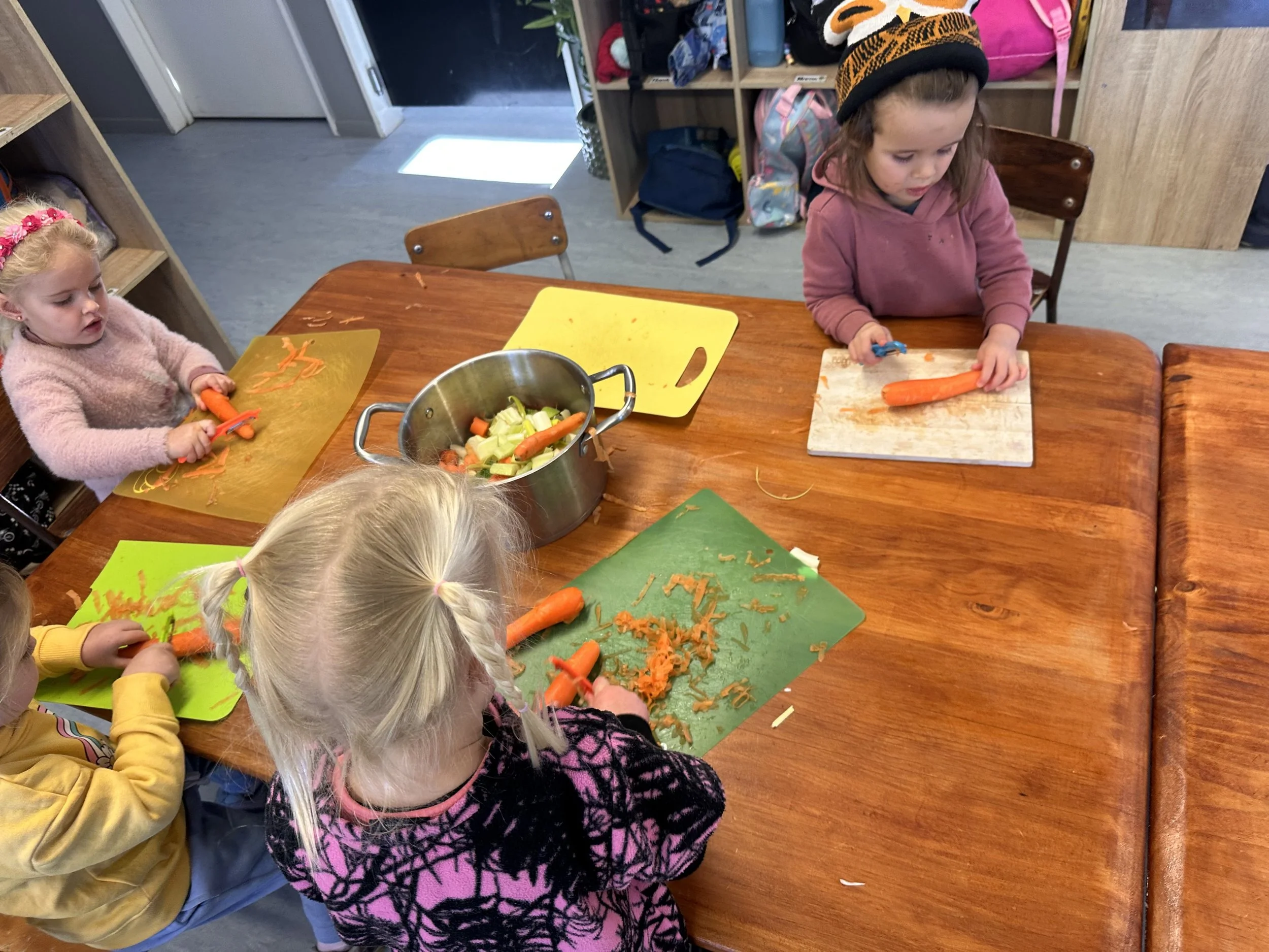 Children sitting at a wooden table peeling and chopping carrots and vegetables, with a pot of vegetables on the table.