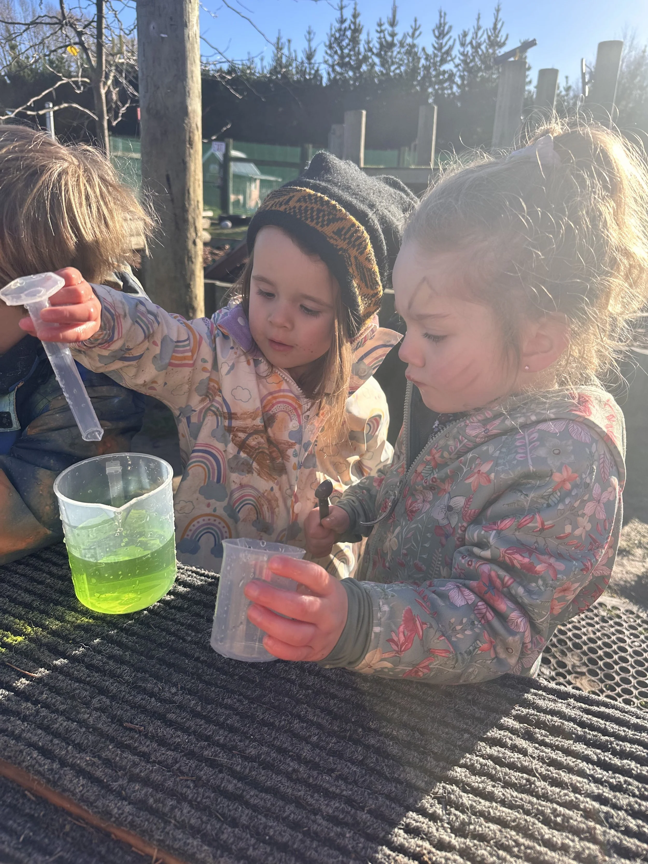 Three young girls outdoors in sunlight, involved in a science activity, with containers of colorful liquids on a black textured surface.