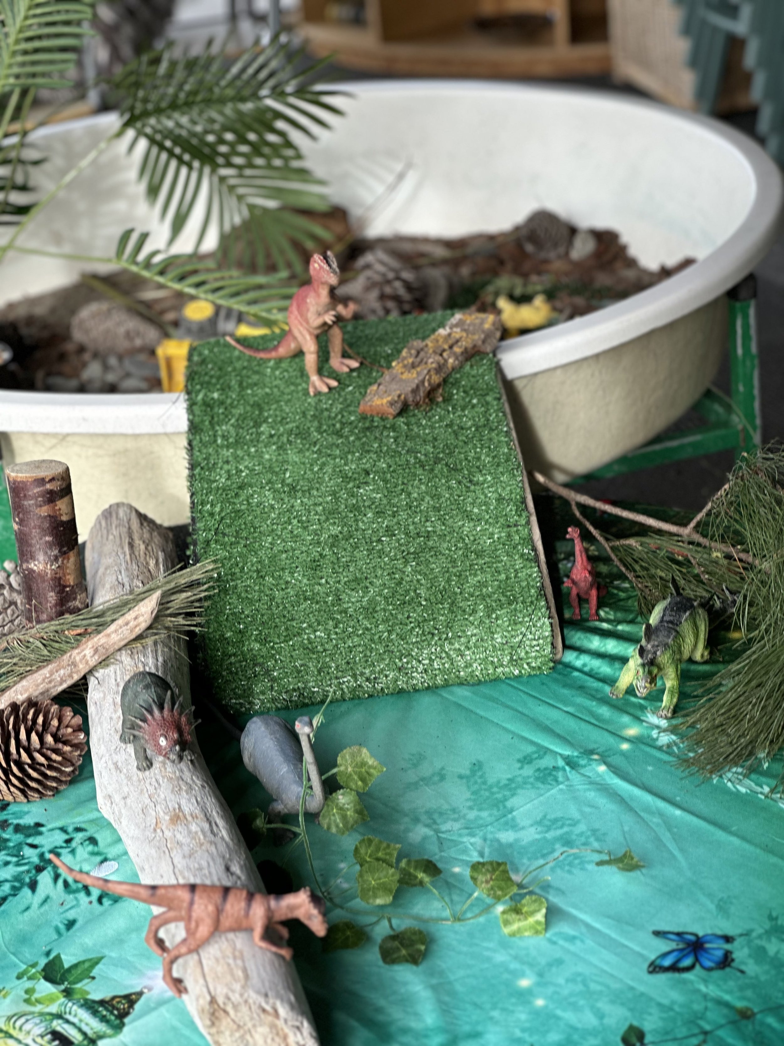 A small toy dinosaur on a green platform with a textured surface, surrounded by various plants, rocks, and animal figurines, set up as a miniature scene.