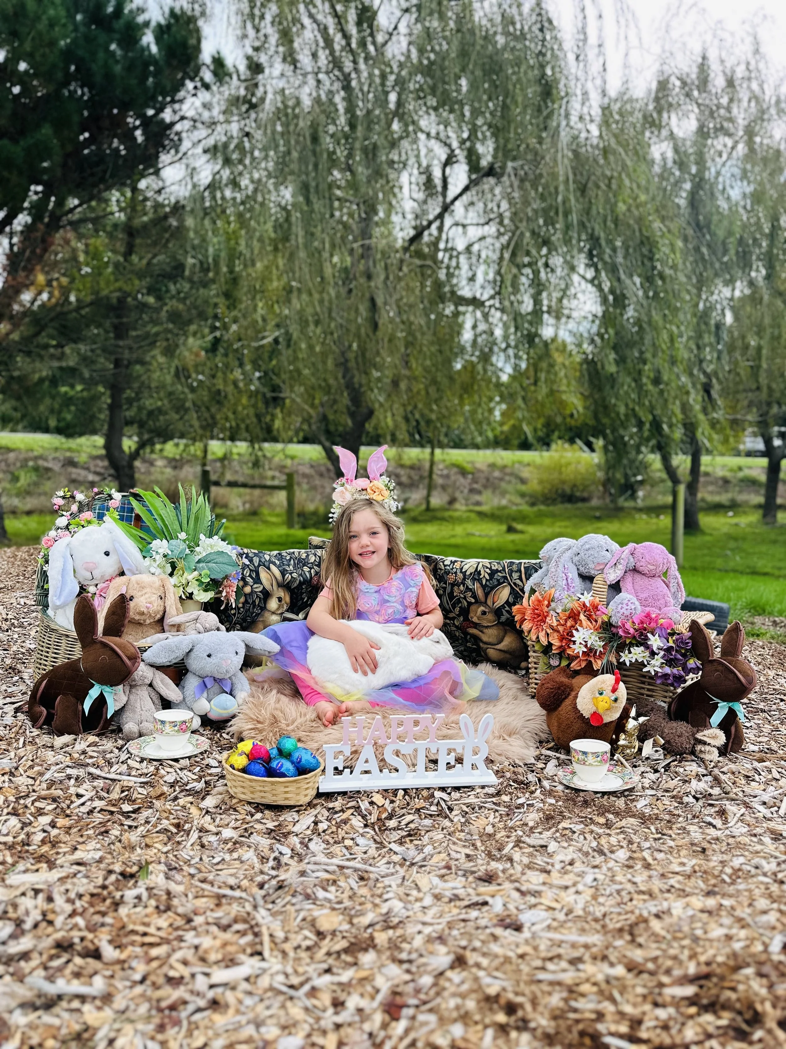 A young girl in a pastel dress, wearing a bunny ears headband, sits on a cozy rug surrounded by Easter-themed decorations and plush bunny stuffed animals, with a sign reading 'HAPPY EASTER' and a basket of colorful Easter eggs outside, outdoors in a 