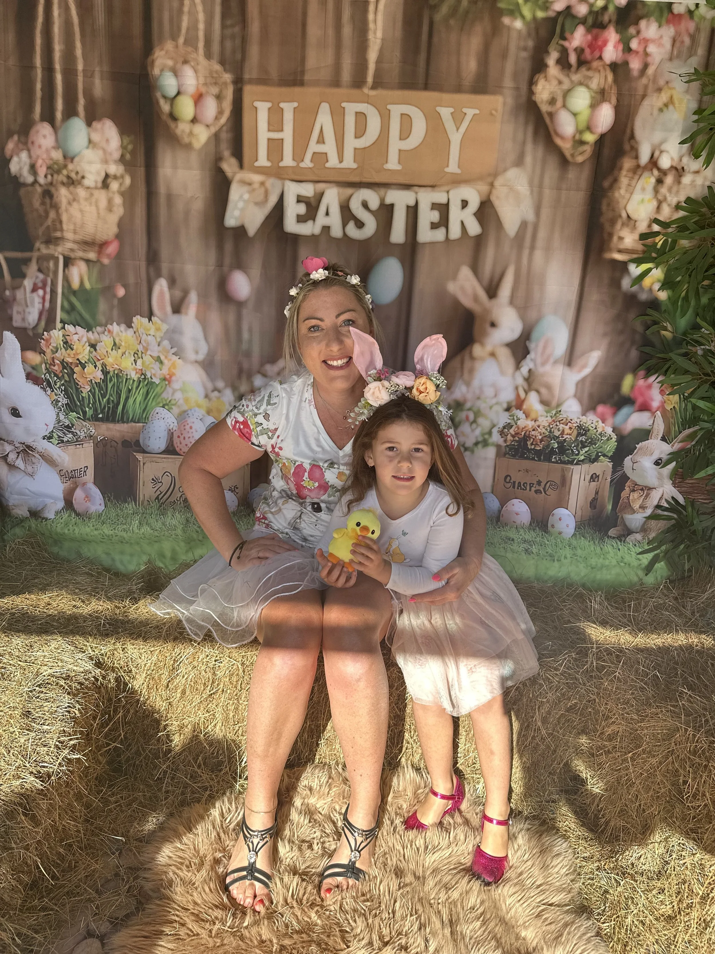 Two women, one adult and one child, sitting on a faux fur rug in front of a festive Easter-themed backdrop. The backdrop features baskets of pastel-colored eggs, bunny decorations, and a sign that says "HAPPY EASTER." The adult woman is wearing a flo