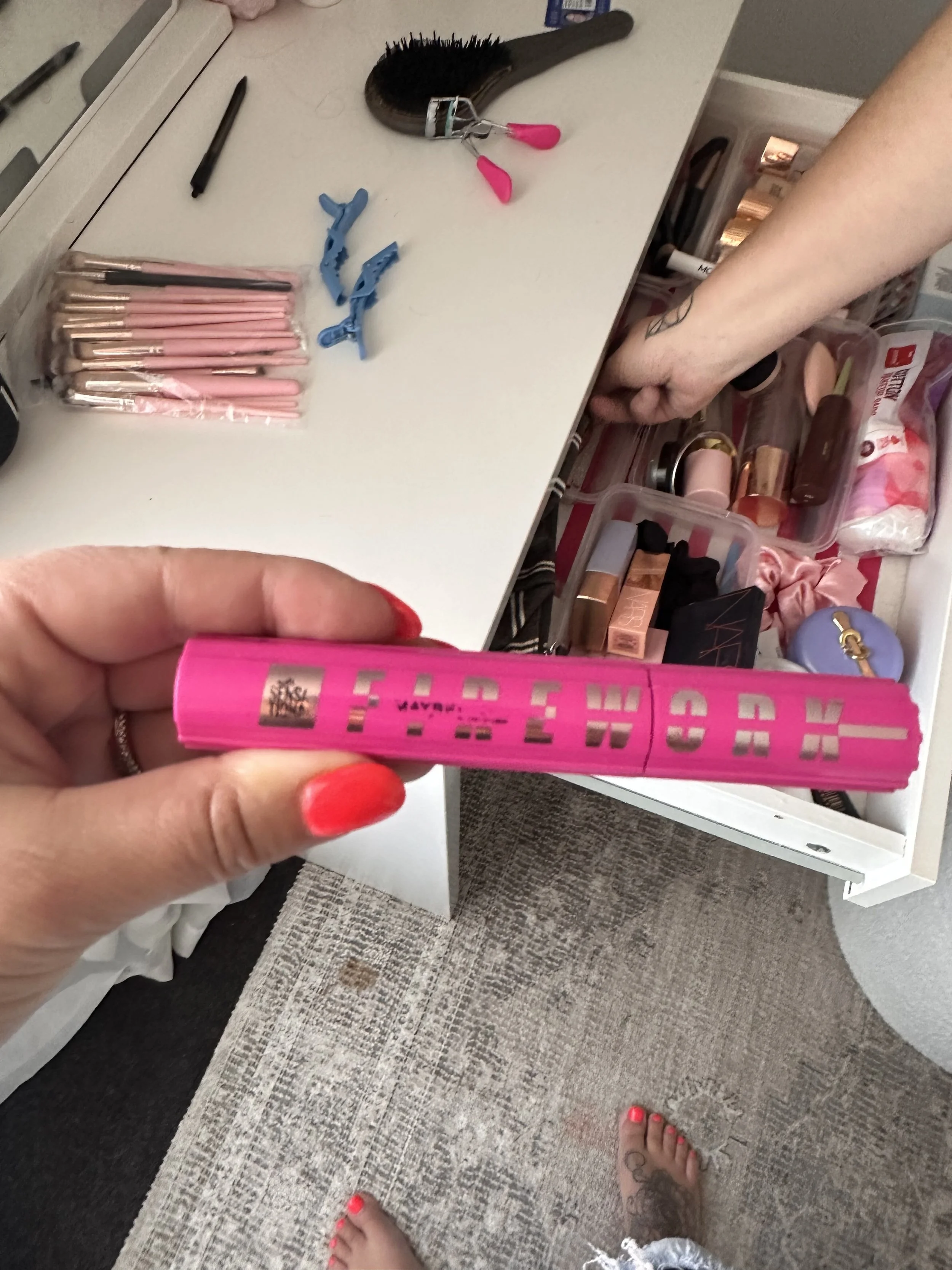 A person's hand holding a pink lipstick tube with gold accents. In the background, there is a white surface with various makeup brushes in pink cases, hairbrush, makeup items, and a hand reaching into a container of makeup products. The person has th