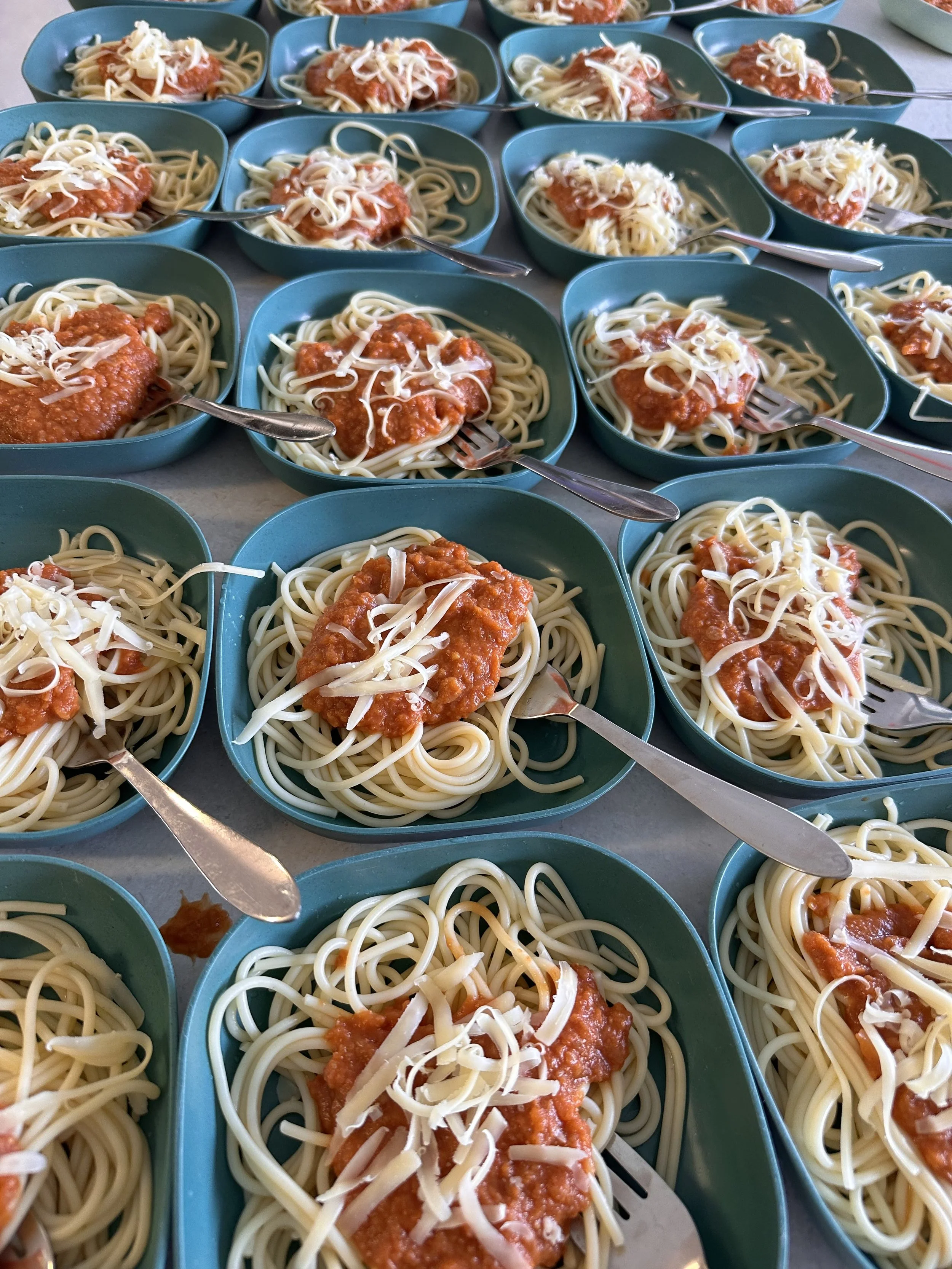 Multiple teal-colored bowls filled with spaghetti noodles topped with marinara sauce and shredded cheese, with metal forks resting inside each bowl.