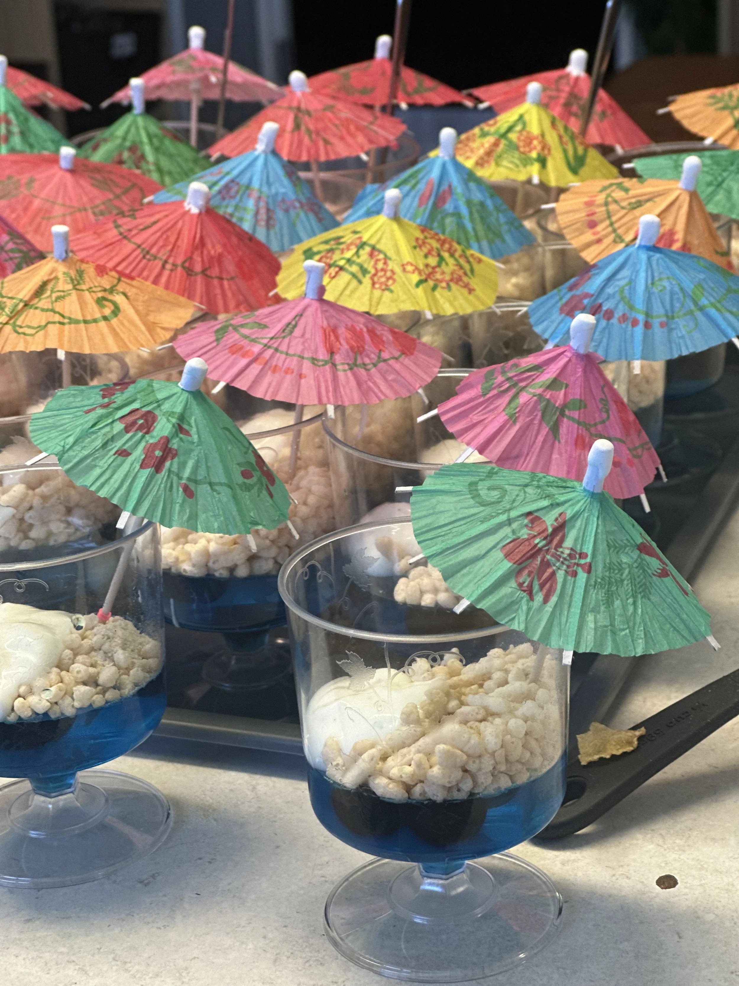 Multiple plastic cups filled with ice cream, decorated with small colorful paper umbrellas, arranged on a counter.