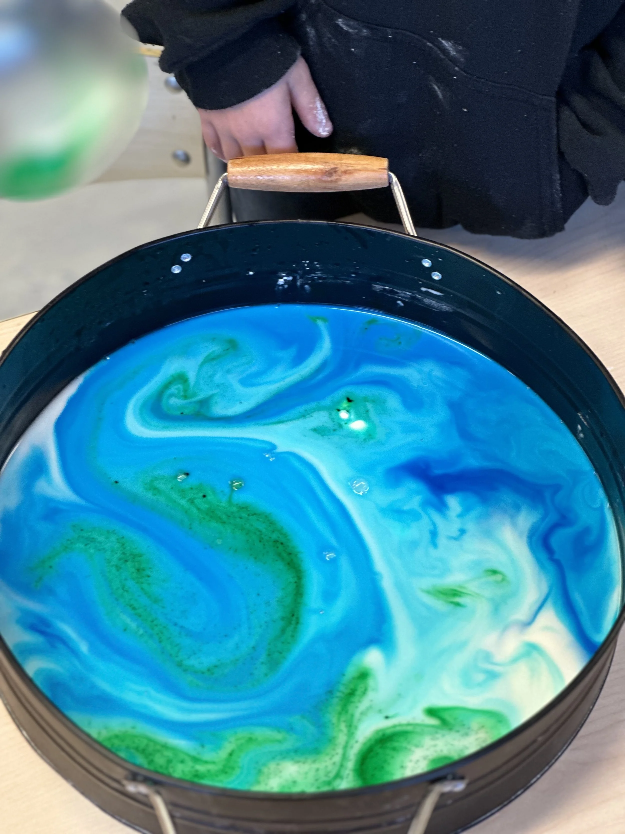 Top-down view of a black pot with a swirling mixture of blue, green, and white colors inside, placed on a light-colored surface.