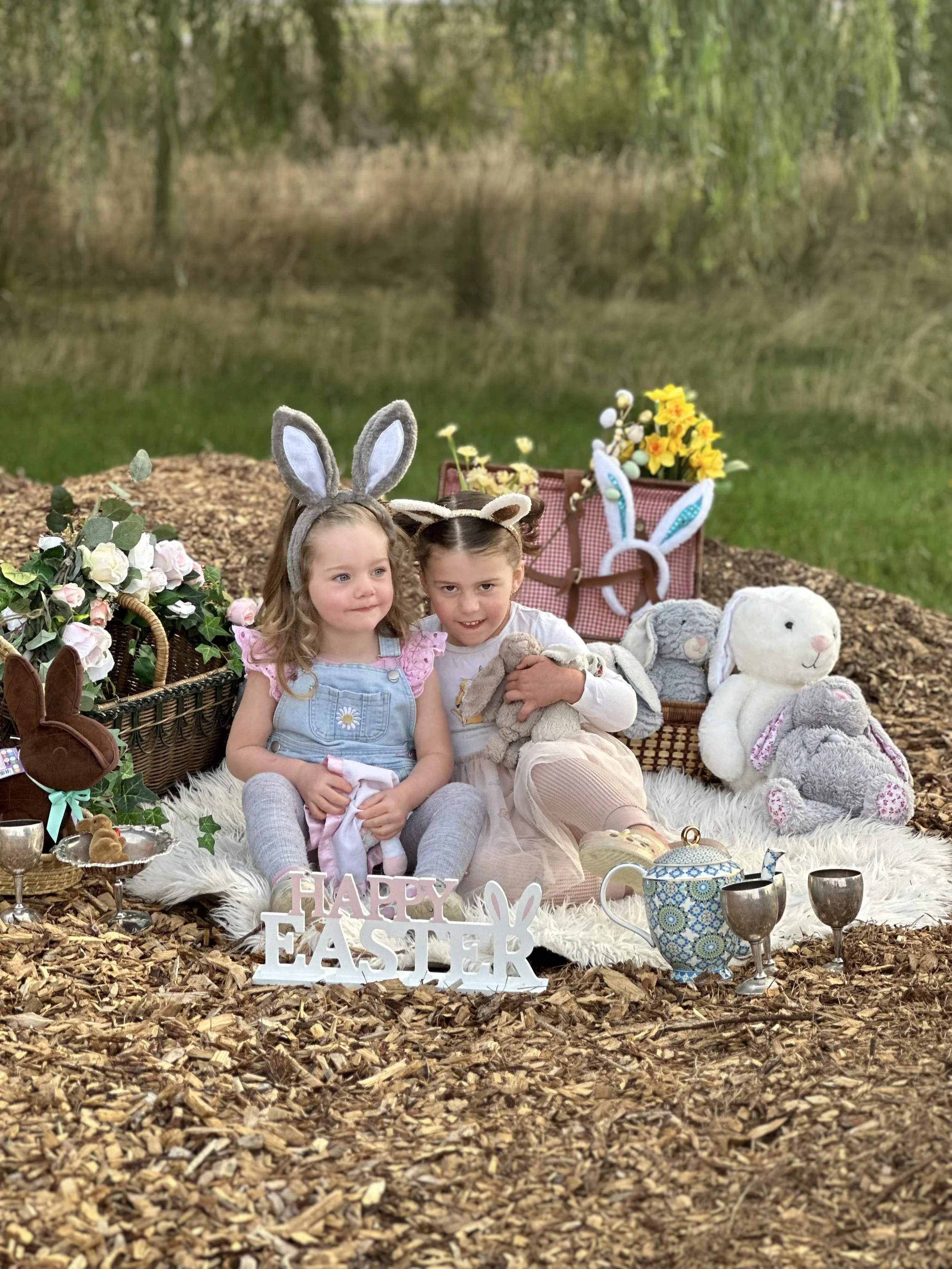 Two young girls with bunny ear headbands sitting on a white furry rug surrounded by Easter decorations including stuffed bunnies, flowers, a sign that reads 'Happy Easter,' a teapot, and cups, outdoor woodland setting.