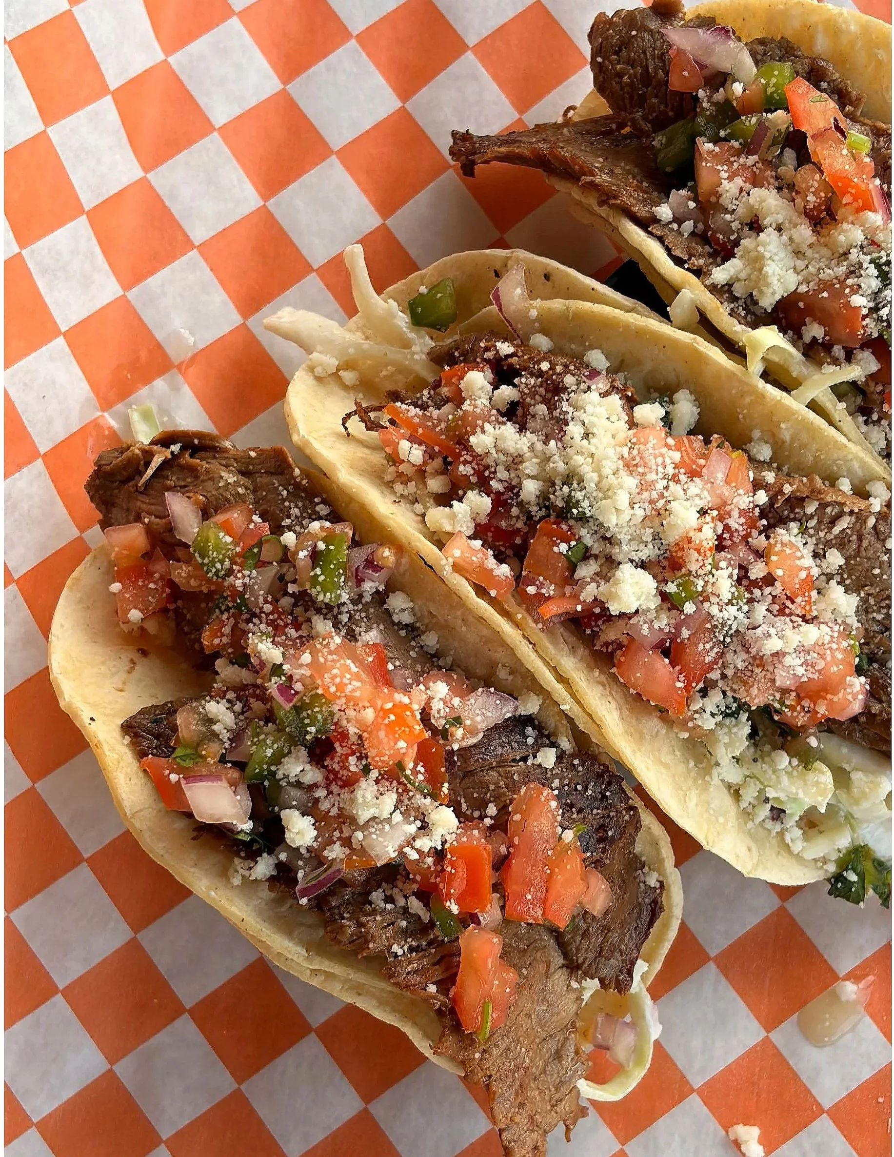 Close-up of three tacos with beef, diced tomatoes, chopped onions, chopped green peppers, shredded cheese, and cilantro on orange and white checkered paper.