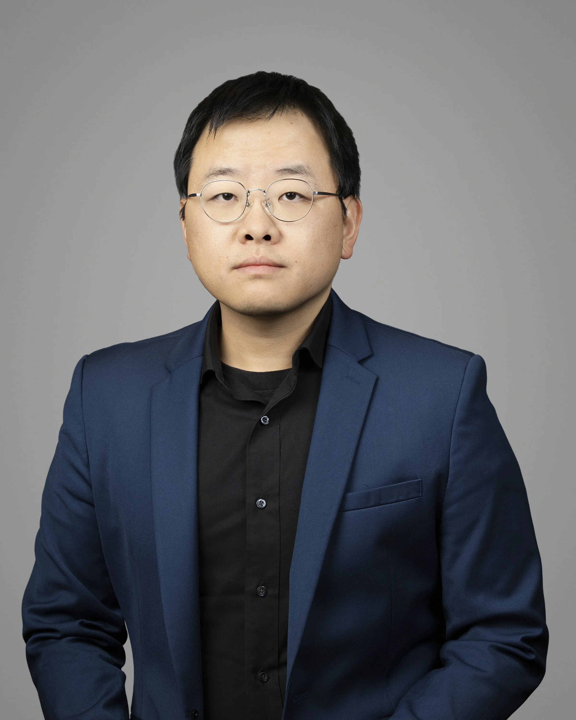 A professional portrait of an Asian man with short black hair, wearing round glasses, a black shirt, and a dark blue blazer, against a plain gray background.