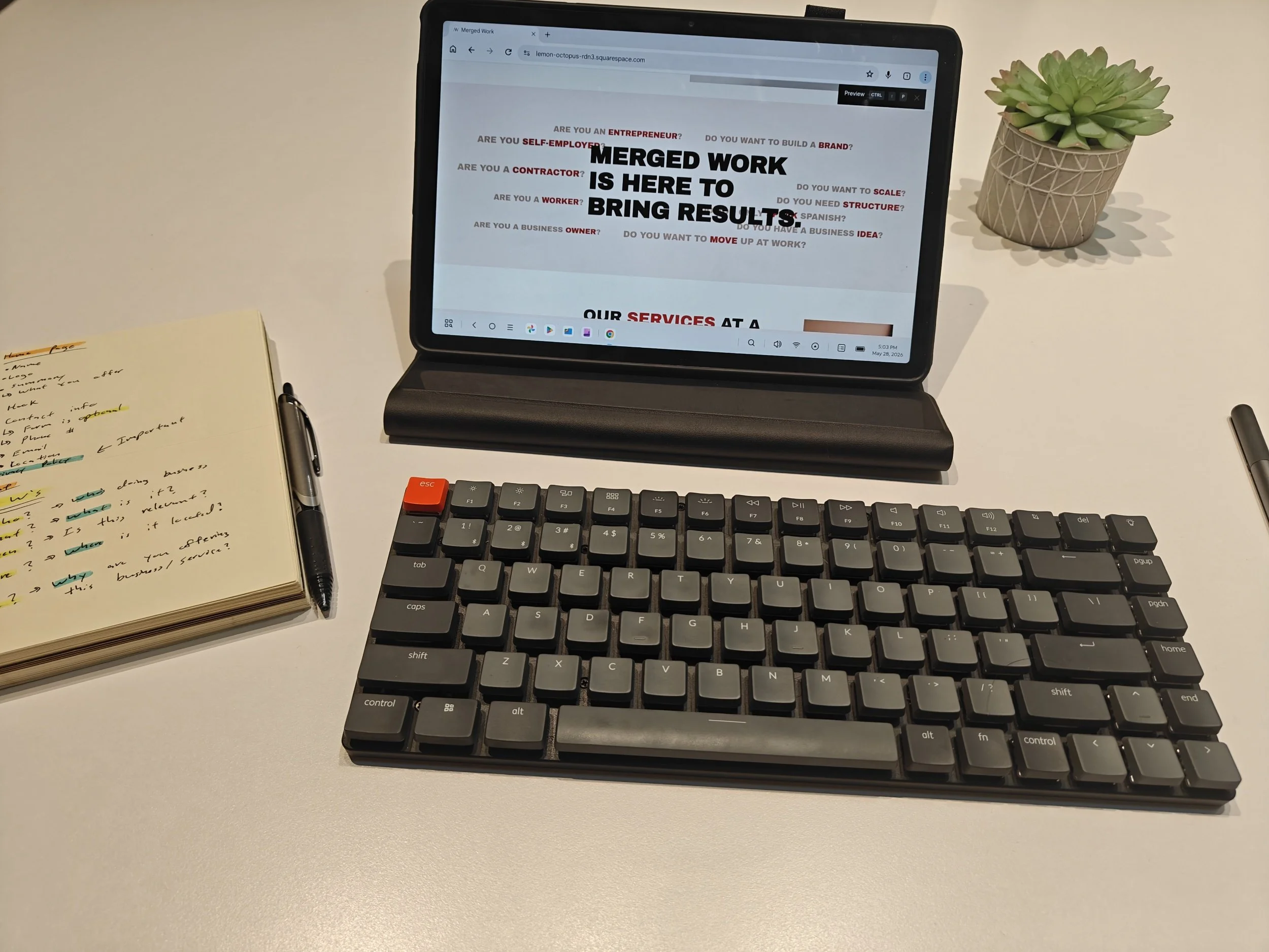 A tablet on a stand displaying a webpage with the headline 'Merged Work is Here to Bring Results,' surrounded by questions about entrepreneurship, self-employment, contracting, working, scaling, branding, structure, language, ideas, business ownership, and moving up at work. A black keyboard is in front of the tablet. To the left, there is a yellow notepad with handwritten notes and a pen. To the right, there is a potted succulent plant. The tablet is on a white desk with a plain light-colored wall in the background.