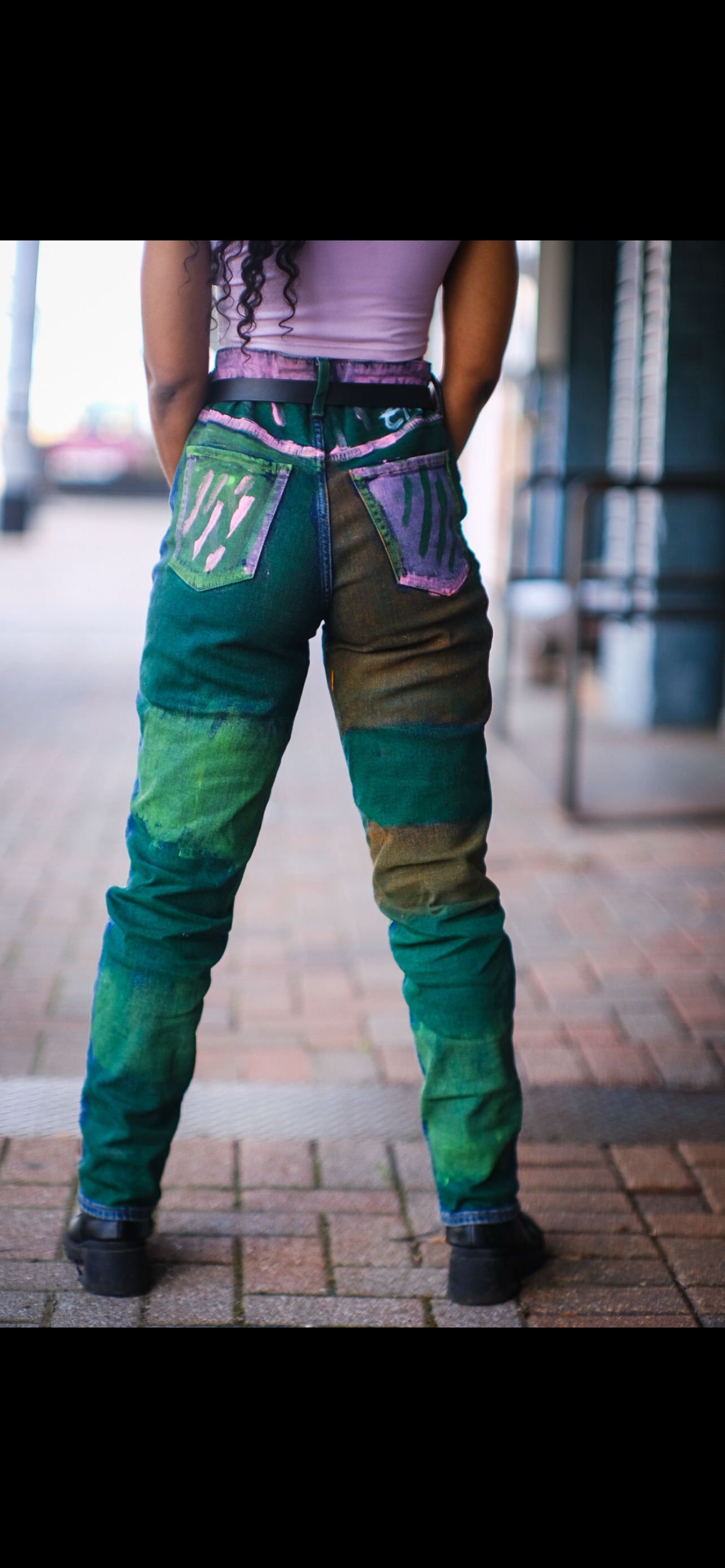 Person wearing colorful, artistic pants with a gradient pattern of green, blue, purple, and brown, standing on a brick sidewalk.