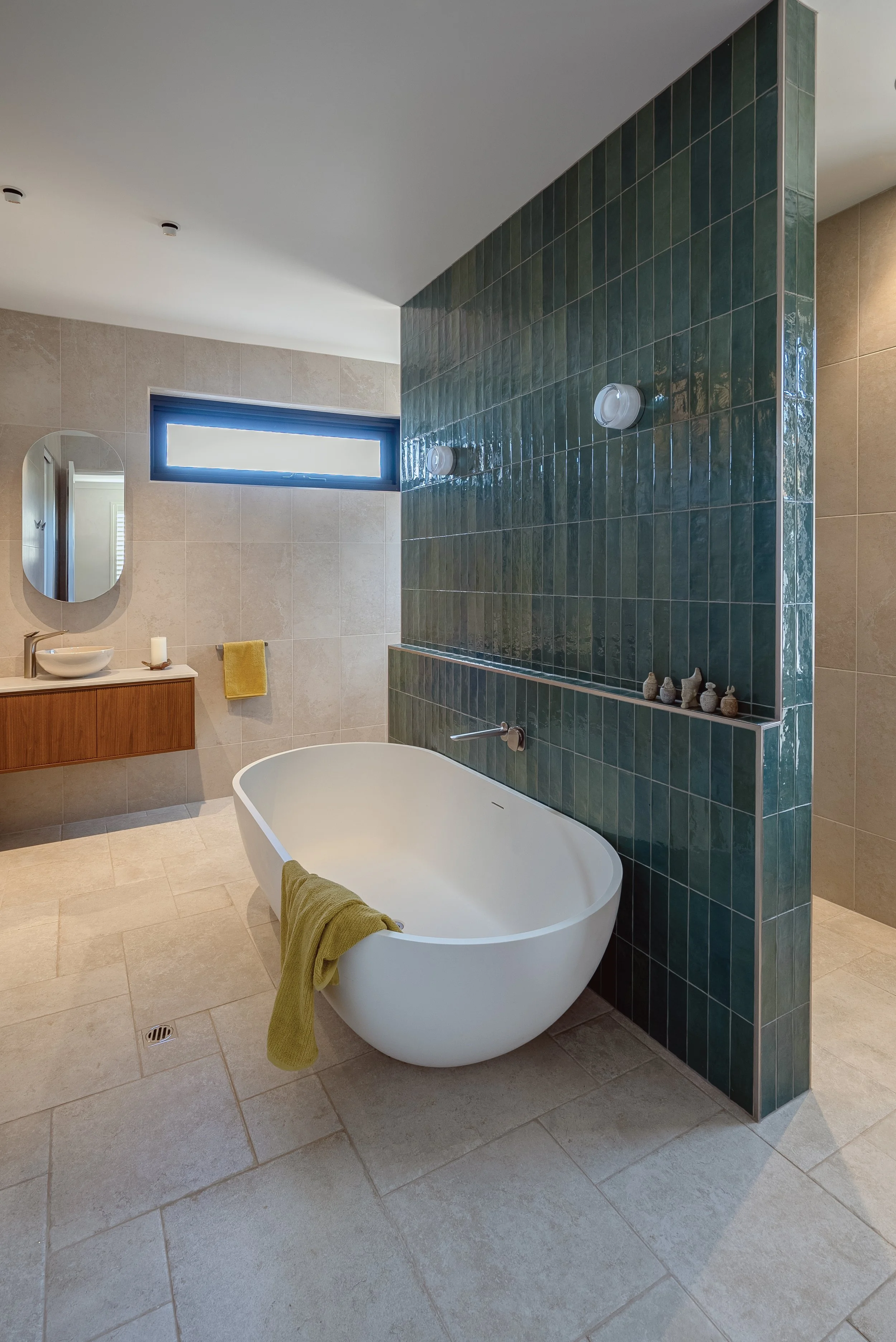 Modern bathroom with a white reconstituted stone freestanding bathtub, green tiled wall, beige tiled floor, small sink with mirror and vase with candle, yellow towel, small decorative objects on ledge, small window, and ceiling lights.