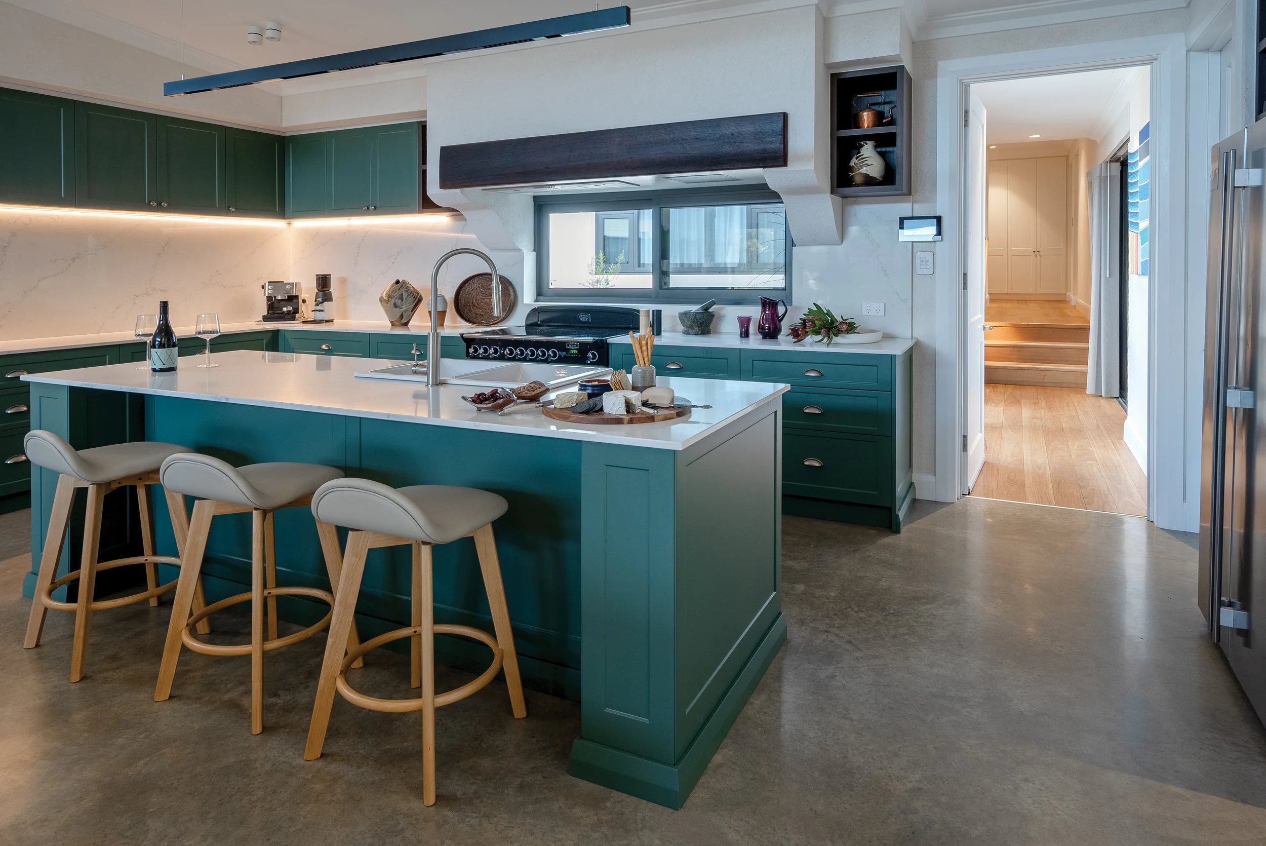 Modern kitchen with green cabinets, white marble countertops, and a kitchen island, decorated with cheese, bread, wine, and vases, with wooden stools and a view of a hallway.