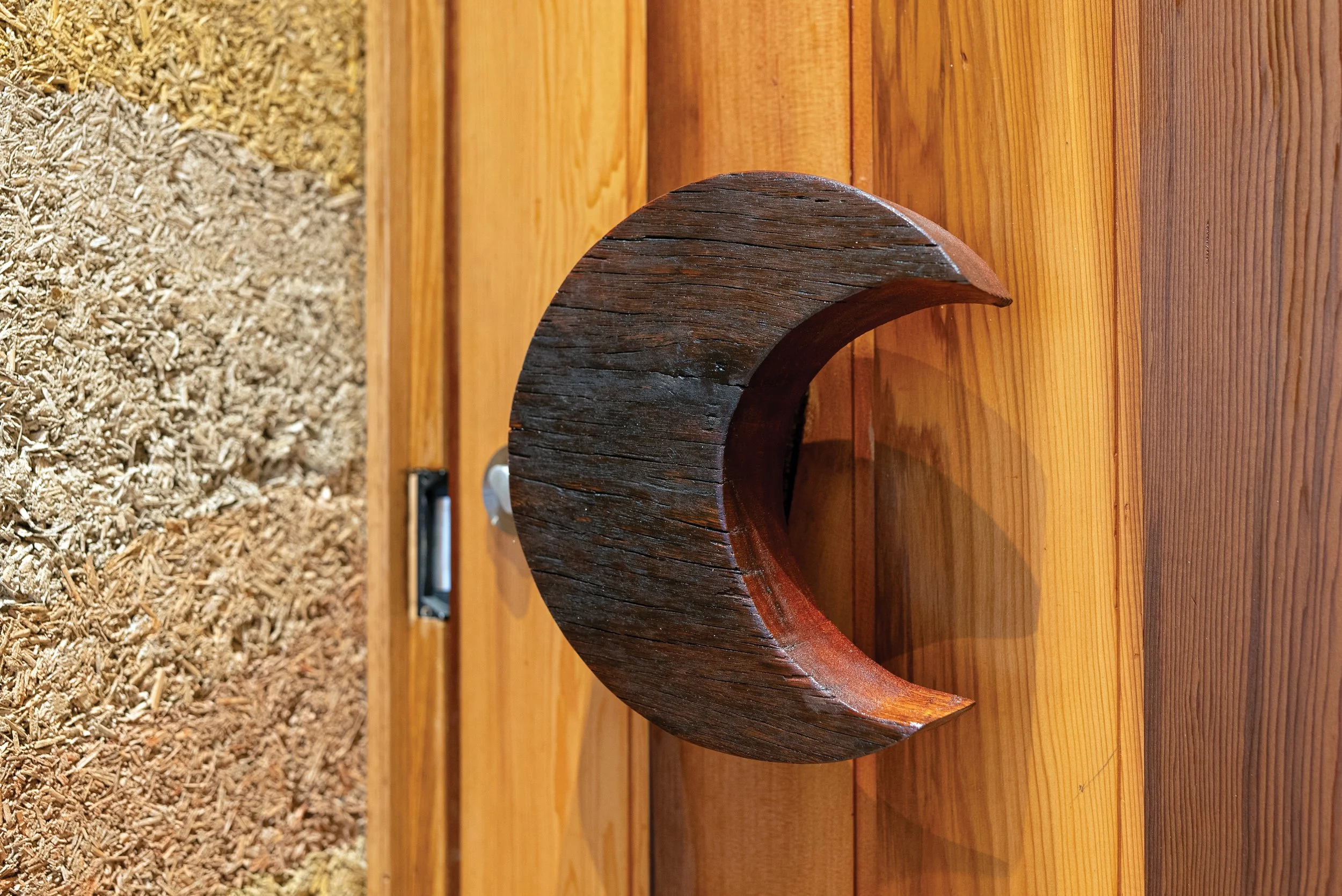 A jarrah crescent door handle on a on a polished Western Red Cedar door, abutting an ochre hempcrete textured wall.