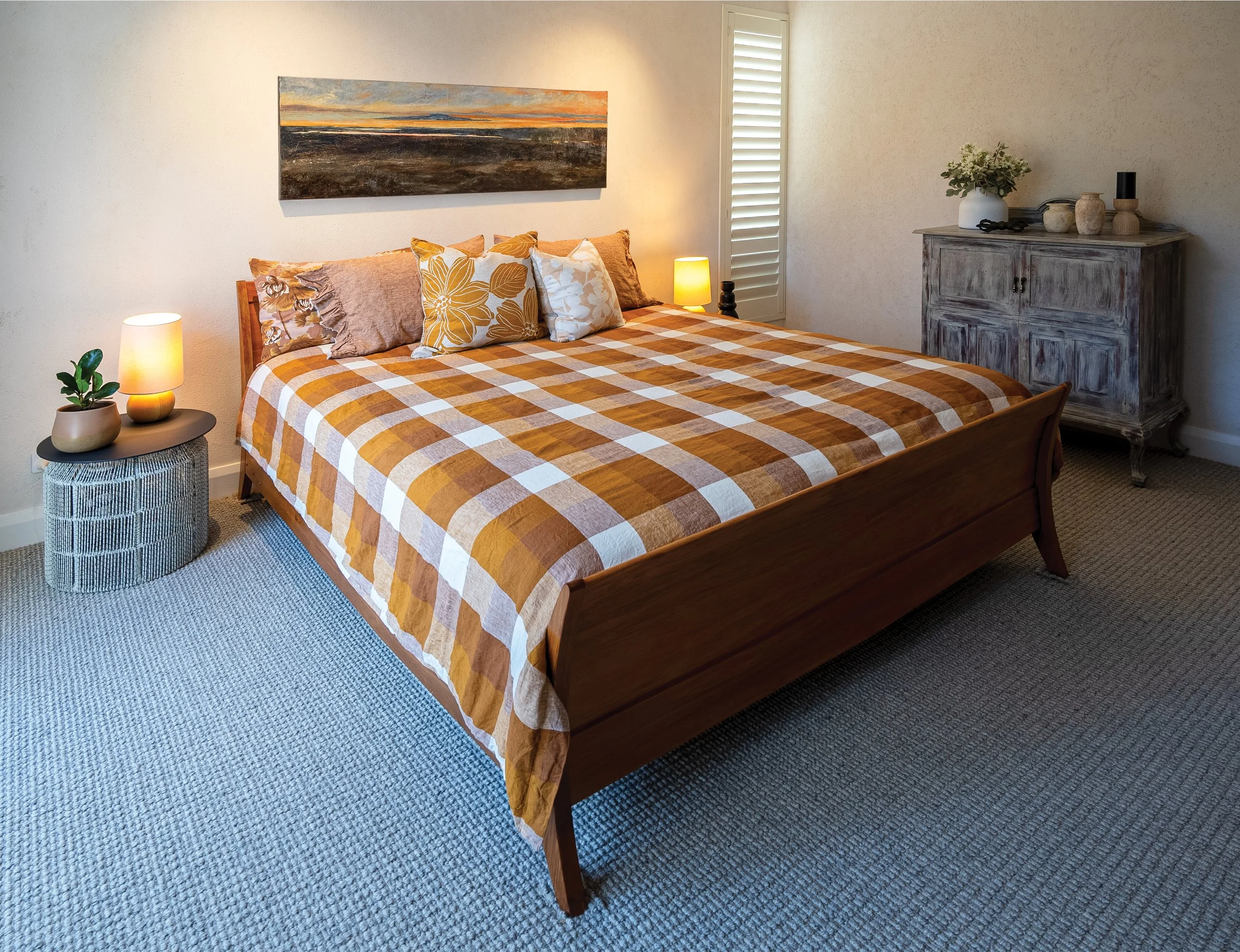 A bedroom with a wooden bed frame, a checkered orange and white bedspread, several pillows, and two bedside lamps. There is a wooden cabinet with decorative vases and a plant, and a window with shutters.