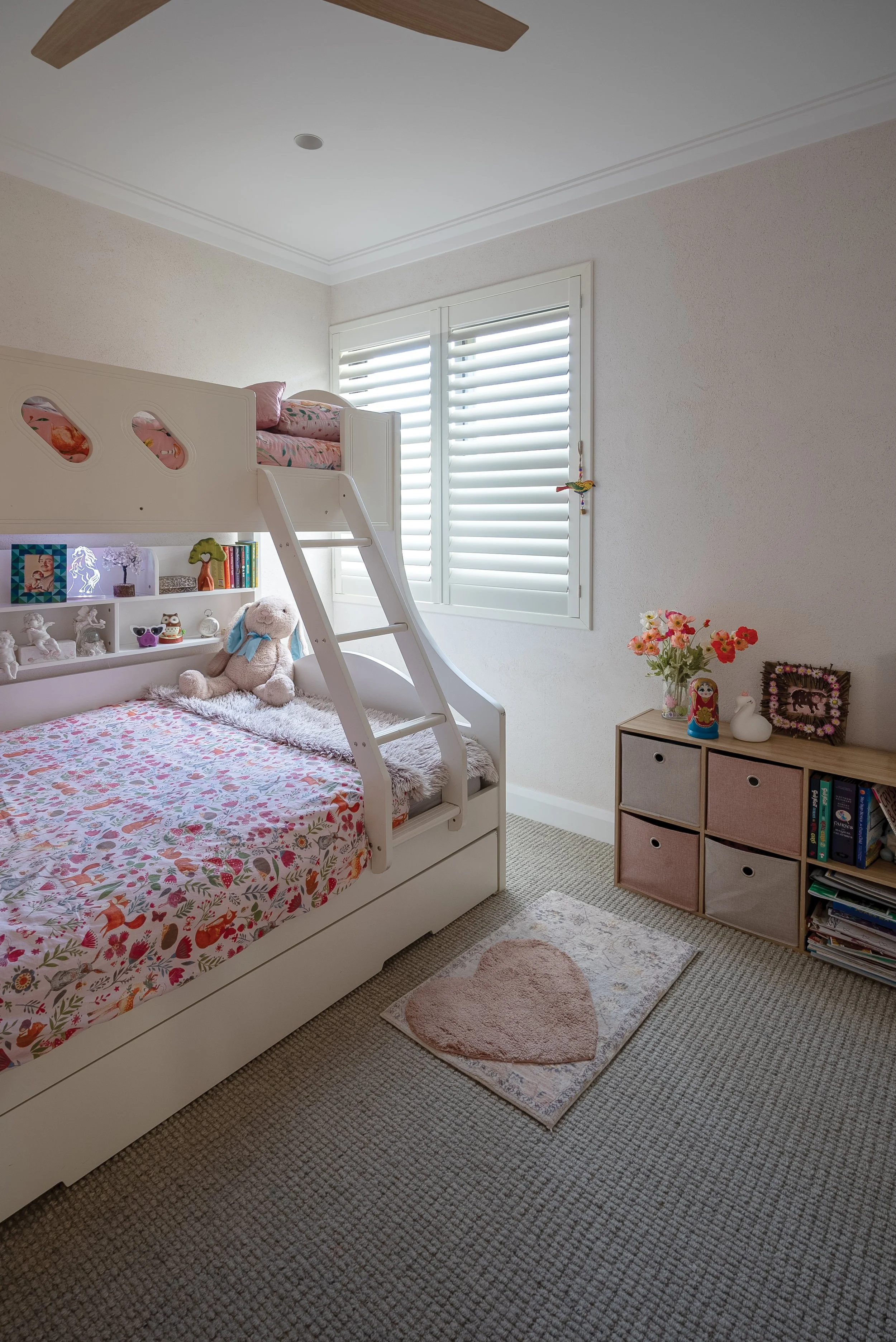 A cozy child's bedroom with a white bunk bed, decorated with pink floral bedding and plush toys. There's a window with white shutters, a small shelving unit with decor and books, and a pink heart-shaped rug on a beige carpeted floor.