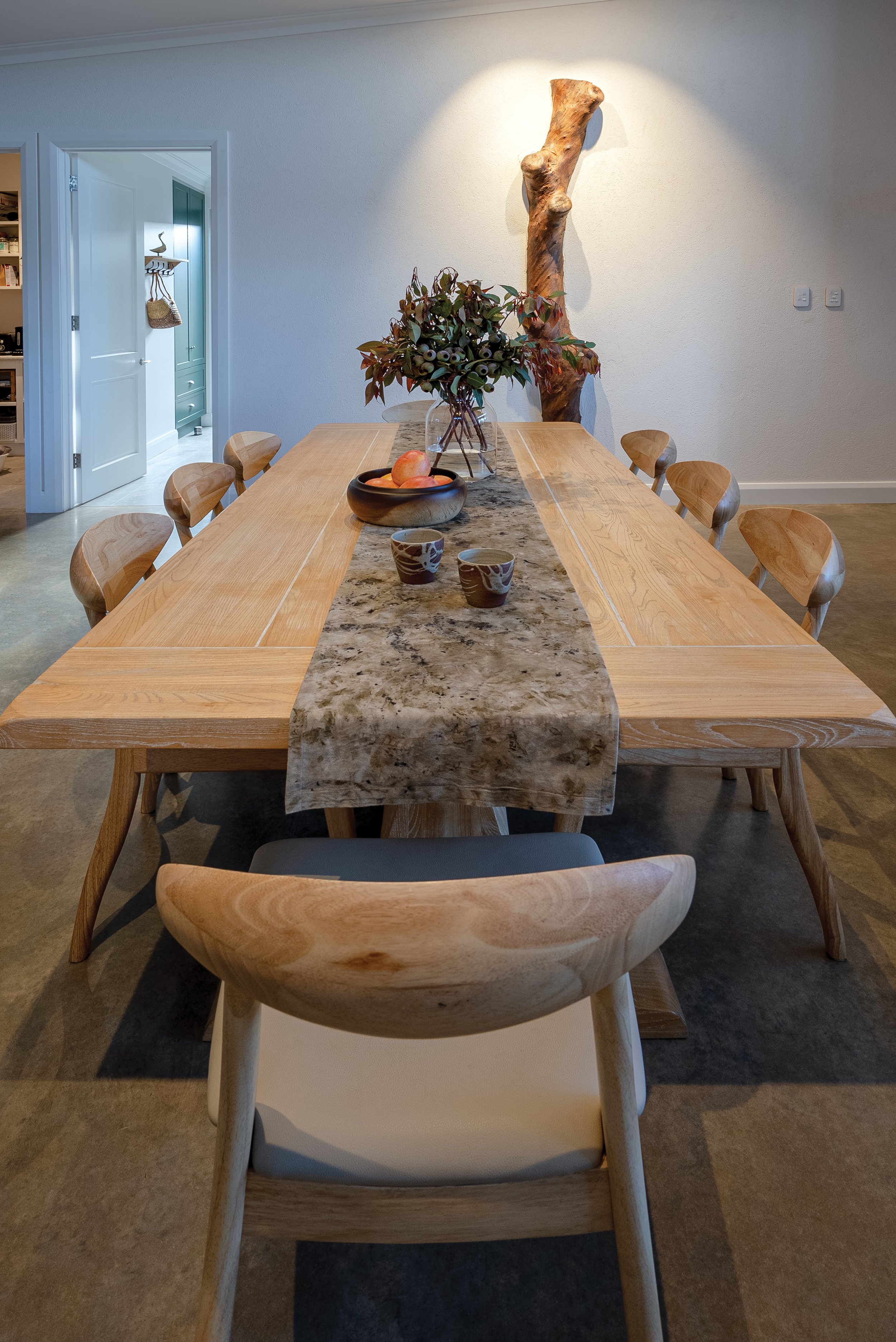 The image features a wooden dining table with 6 chairs.  In the background, a lime rendered hempcrete wall with large abstract timber art illuminated by a spotlight. To the left,  a laundry with built-in shelving with a hanging wicker basket.