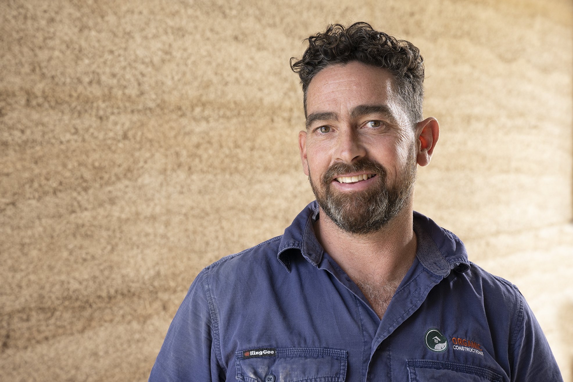 A smiling man in a blue work shirt with a logo, standing in front of a hempcrete wall.