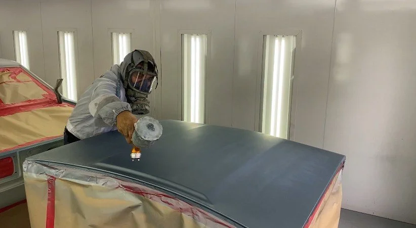 A worker in protective gear spray painting a car hood in a spray booth.