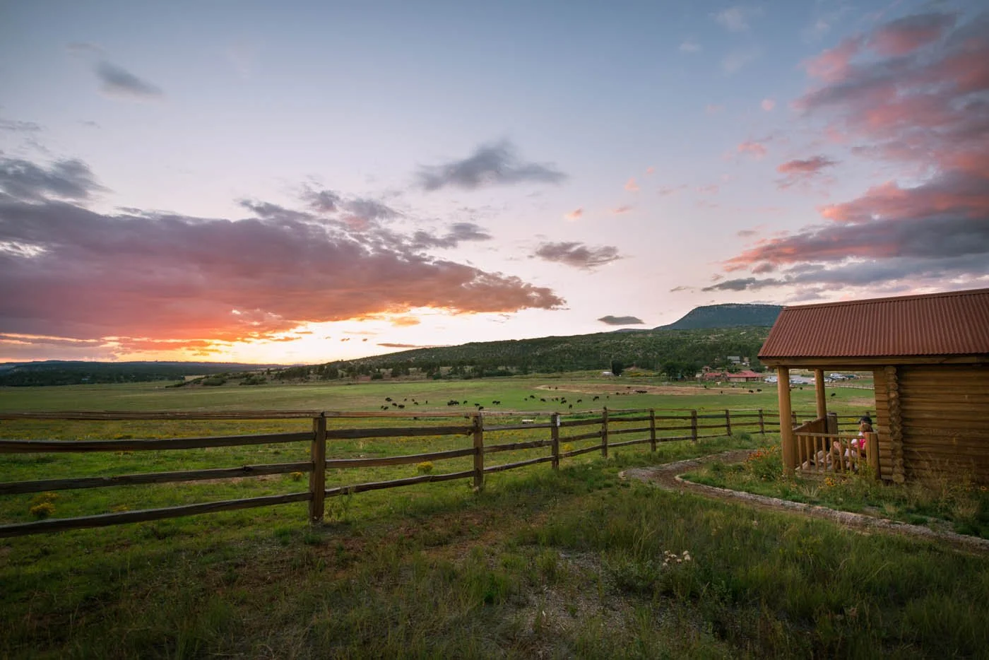 Sunset over a wide green field with a wooden fence, a small log cabin with a red roof, and distant hills with scattered trees.