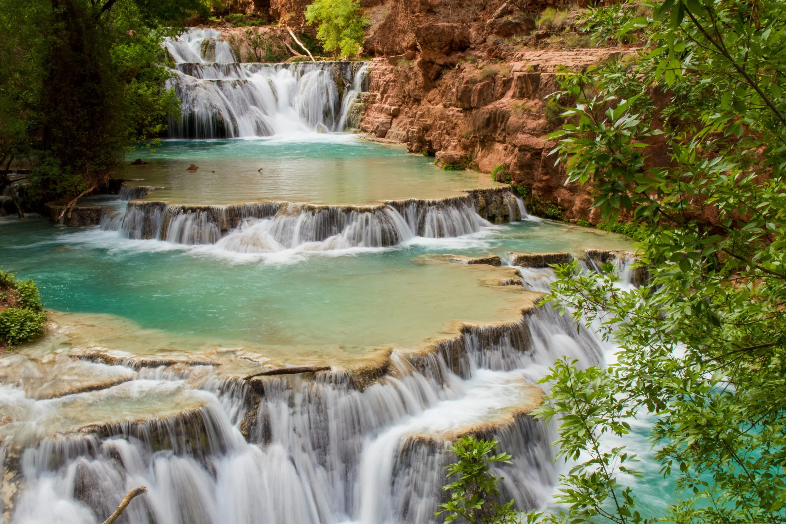 Multiple cascading waterfalls flow into a turquoise pool surrounded by green trees and reddish-brown rocks.