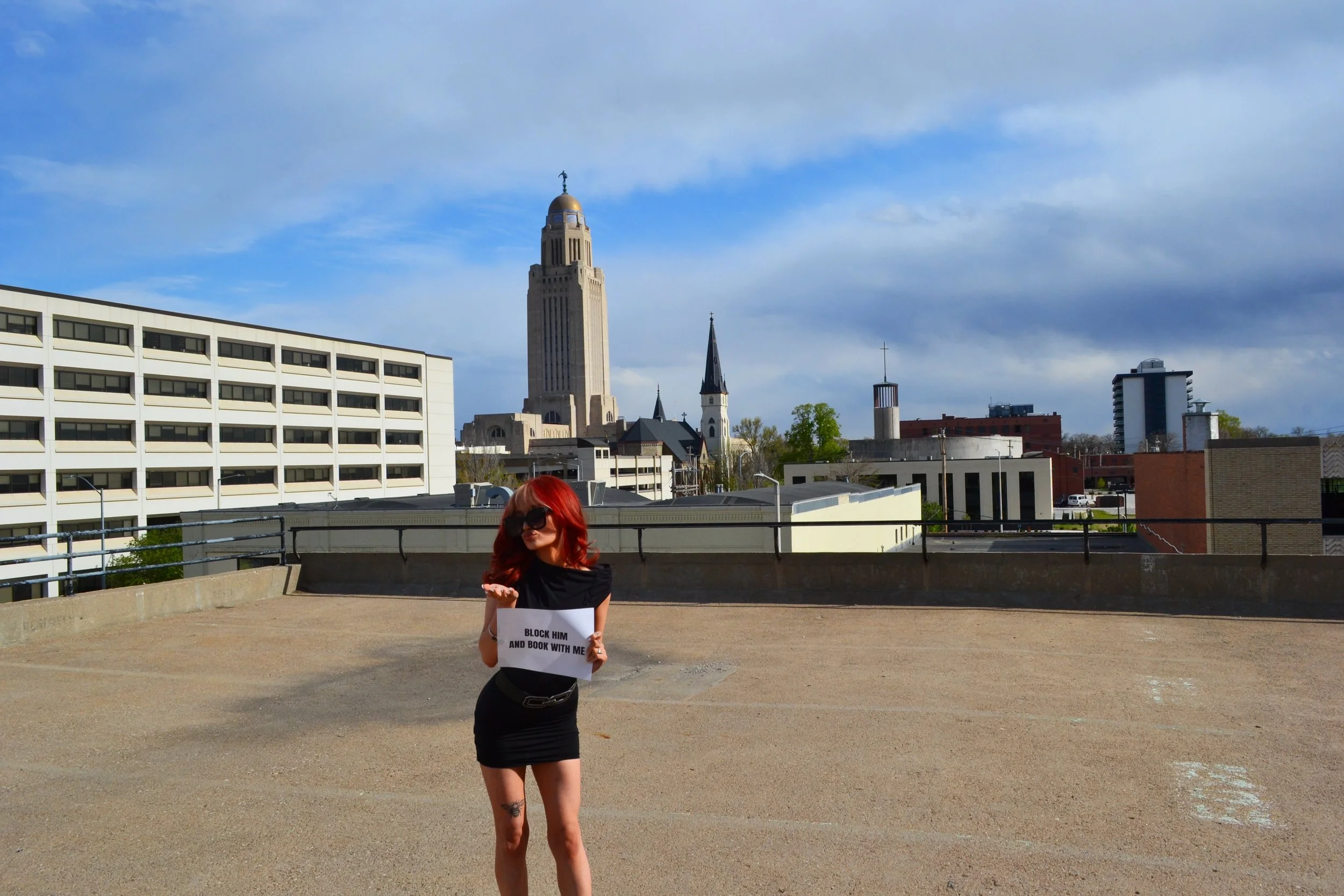 Woman with red hair, sunglasses, and a black dress holding a sign that says 'BLOCK HIM AND BOOK WITH ME' standing on a parking garage roof with city buildings and a tall church steeple and the Capitol building in the background.