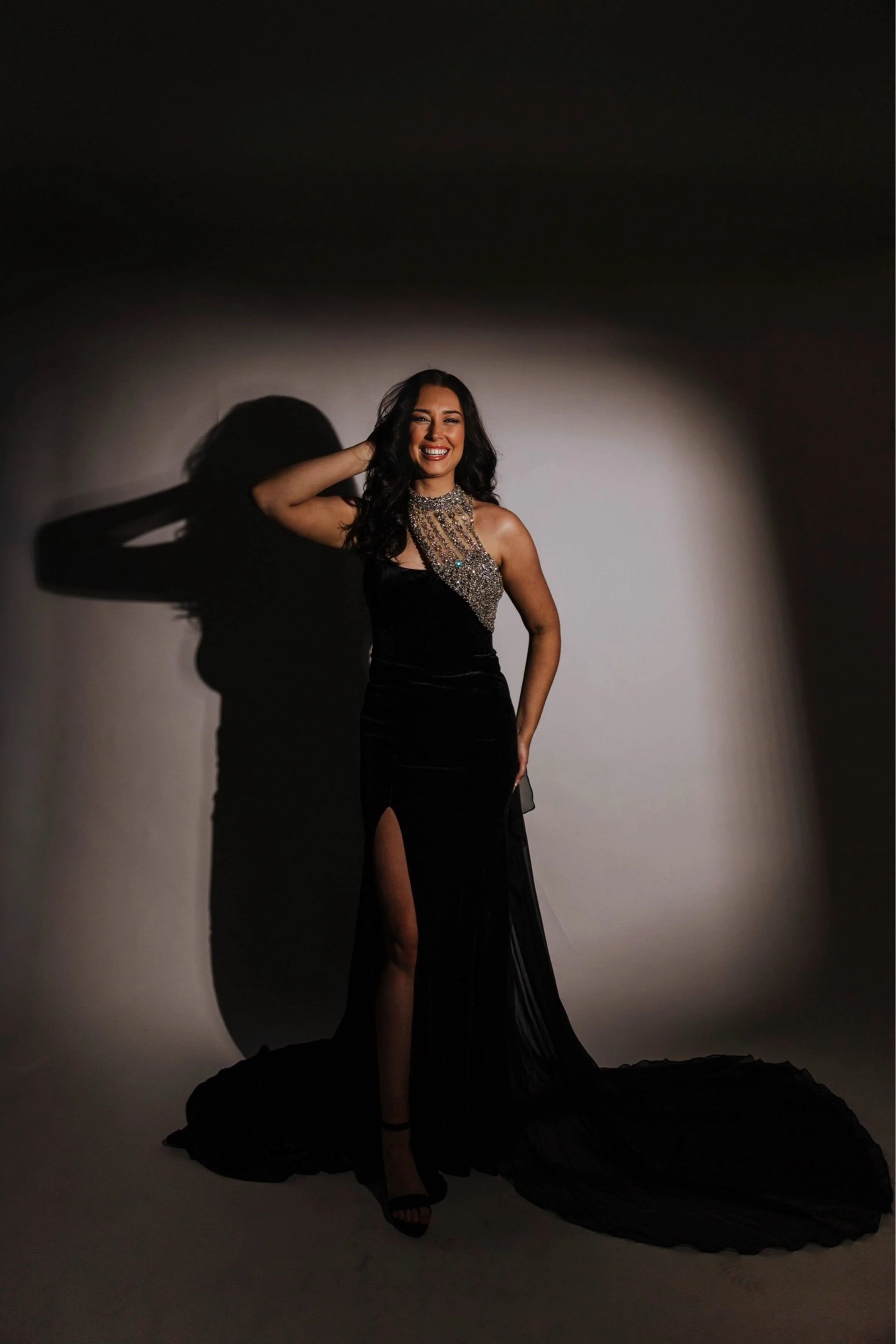 A woman in a long black evening gown with a high slit, smiling, standing against a plain backdrop with a shadow cast on the wall behind her.