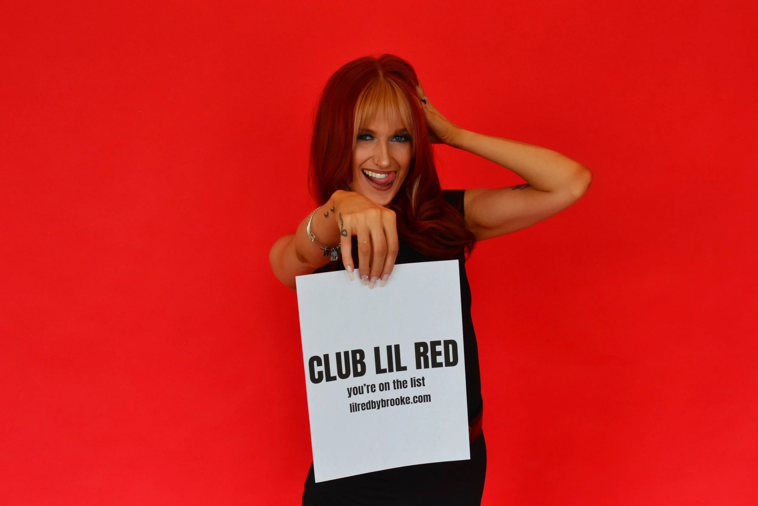 A woman with red hair and tattoos holding a sign that says 'CLUB LIL RED you're on the list lilredbybrooke.com' while smiling and sticking out her tongue against a red background.
