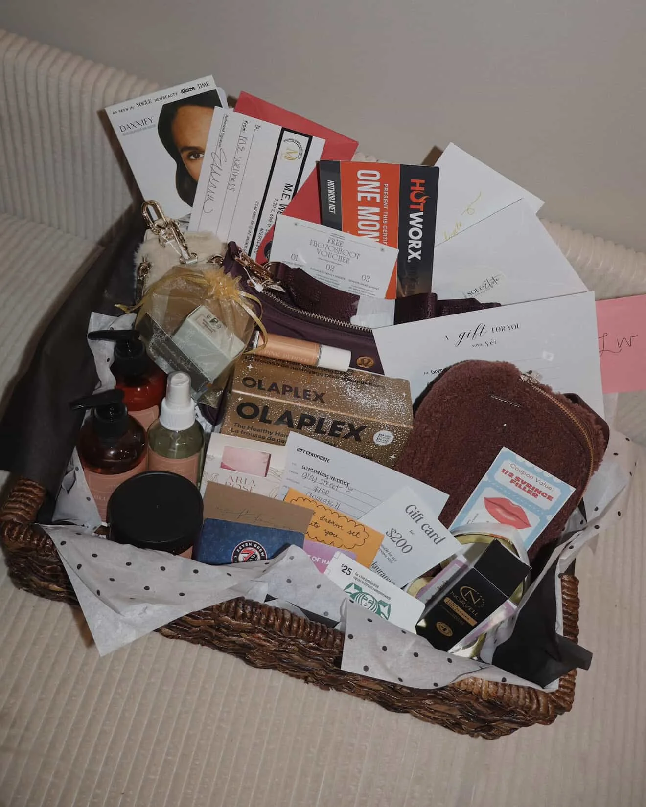 A gift basket containing various items such as skincare products, gift cards, vouchers, a small clutch, and miscellaneous items, lined with white paper with black polka dots.