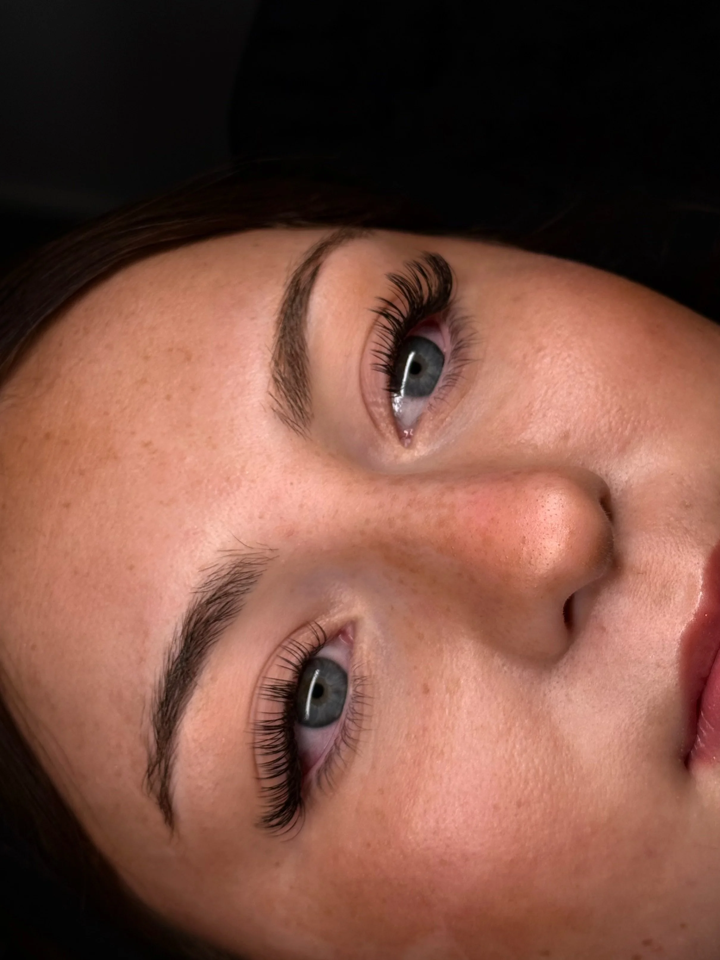 Close-up of a woman's face with clear, light skin, blue eyes, dark eyebrows, and long eyelashes, lying on her side.