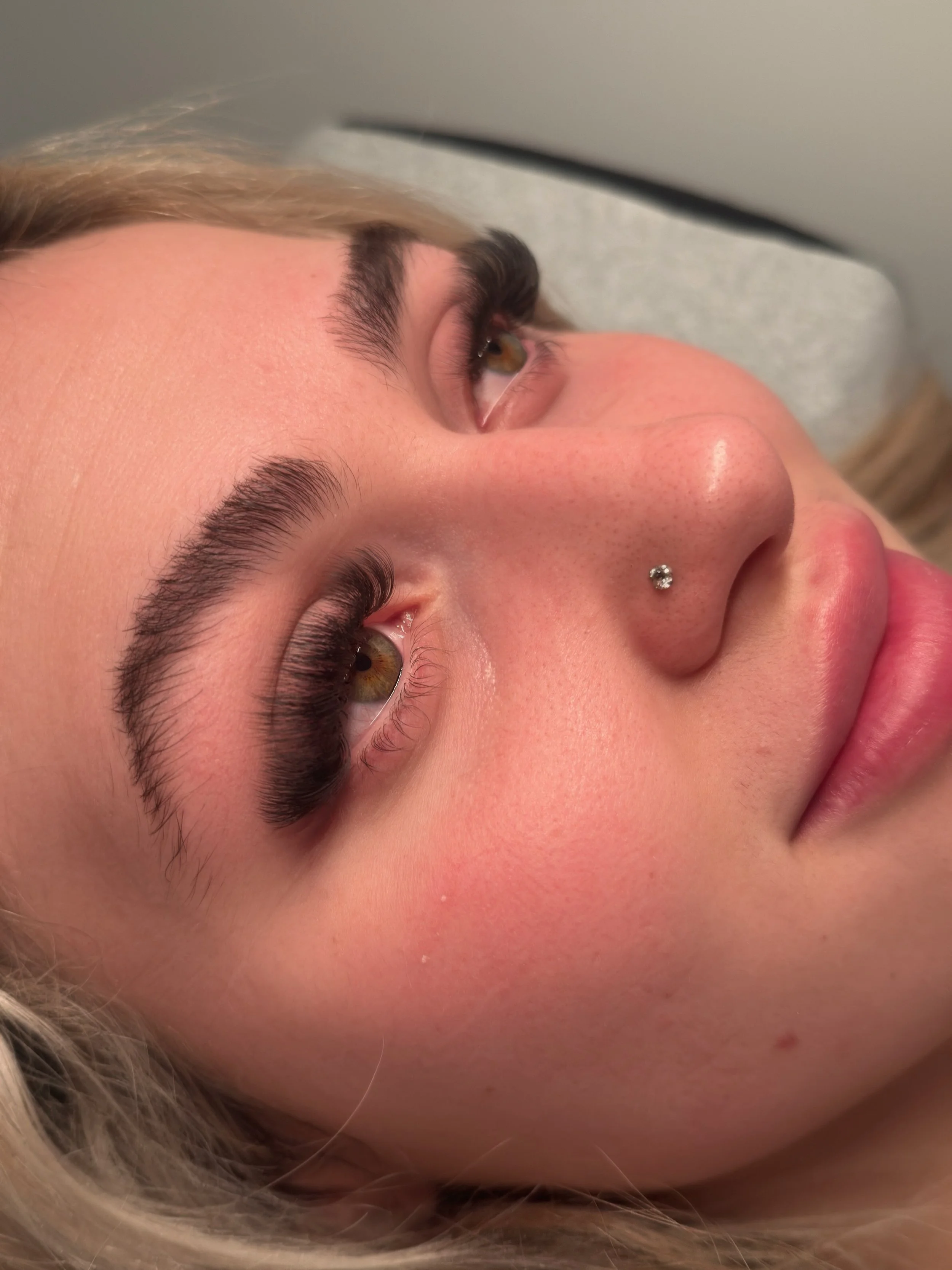 Close-up of a young woman's face lying down, showcasing her eyebrows, eye makeup, nose piercing, and lips.