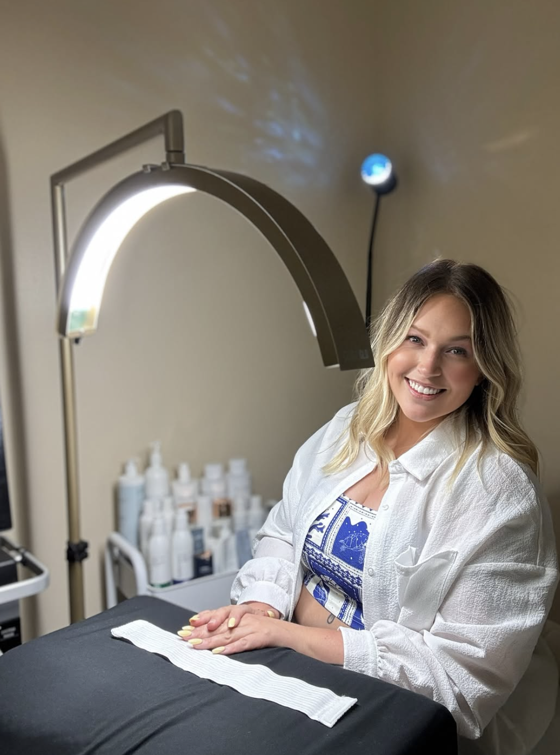 A woman with wavy blonde hair smiling, seated at a skincare or beauty treatment table with a black cloth, with a lamp overhead and various bottles in the background.