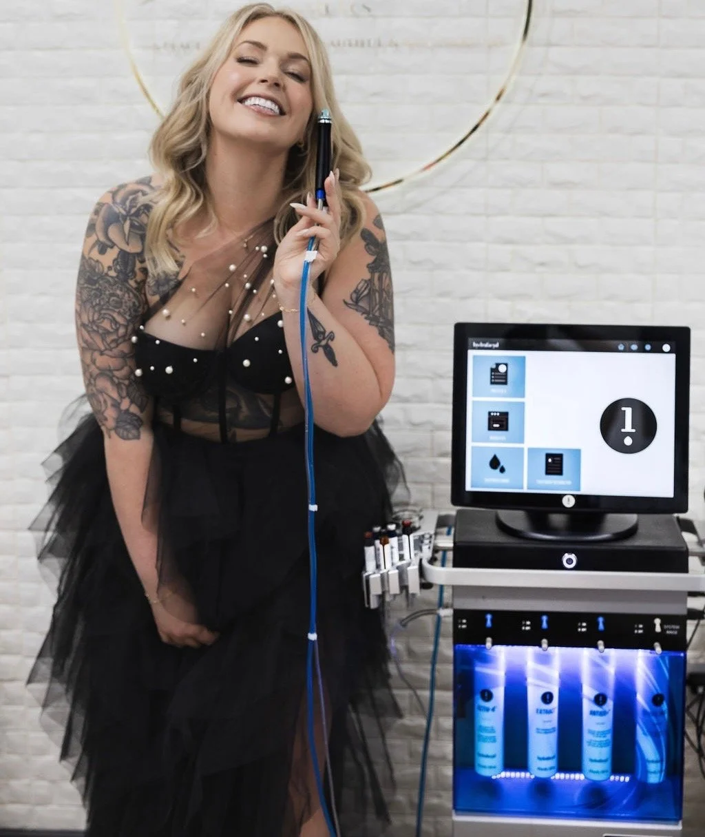 A woman with tattoos on her arms and shoulder, wearing a black tulle dress with pearls, smiling with her eyes closed while holding a microphone connected to equipment. Behind her is a white brick wall and to her right, a medical or beauty machine with tubes and a monitor displaying icons and a number 1.