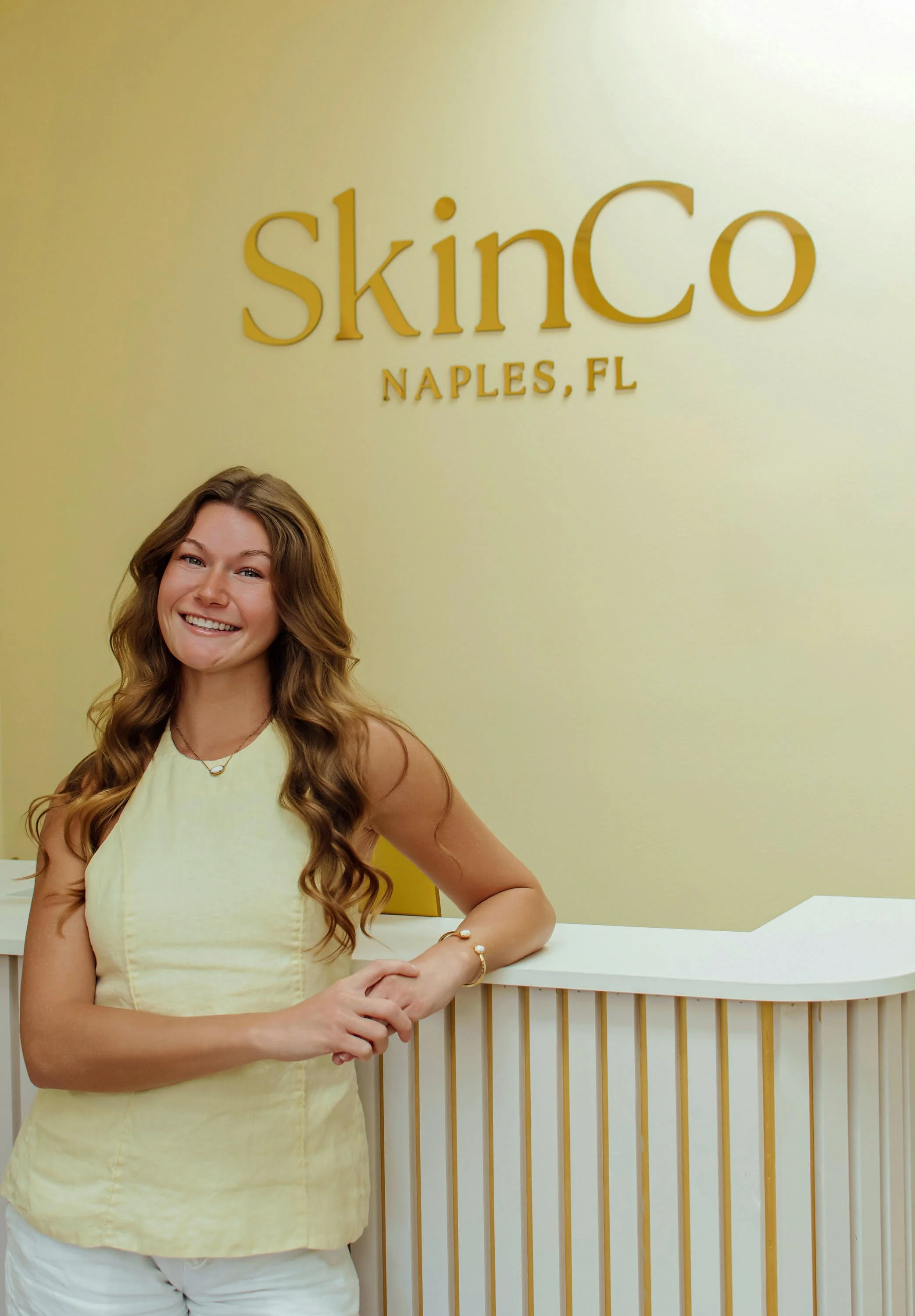 A smiling woman with long, wavy hair wearing a light yellow sleeveless top standing in front of a yellow wall with the words 'skinCo NAPLES, FL' on it. She has jewelry on her wrist and neck, and her left arm is resting on a white counter with a wooden design.