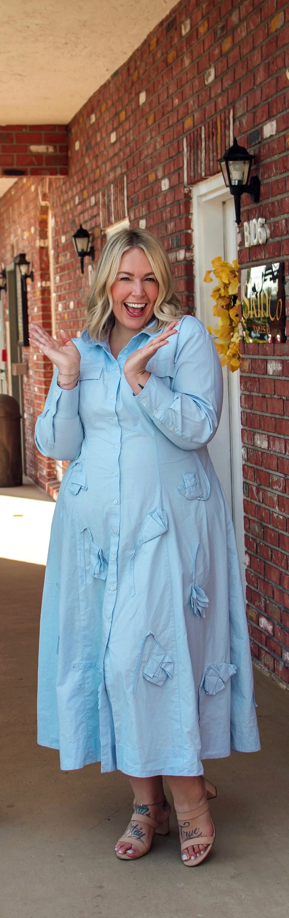 A woman with blonde hair wearing a light blue dress with tied bows, standing outdoors against a brick wall with black lanterns, winking and smiling with hands near her face.