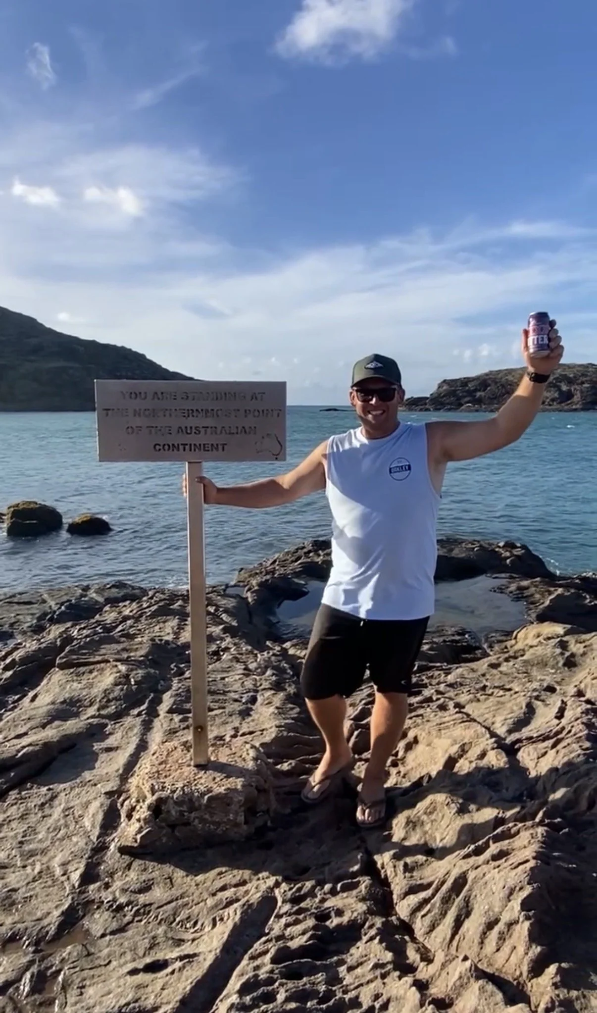 Man standing on rocky shore at the northernmost point of Australia, smiling, holding a sign and a drink, with water and distant islands in the background.