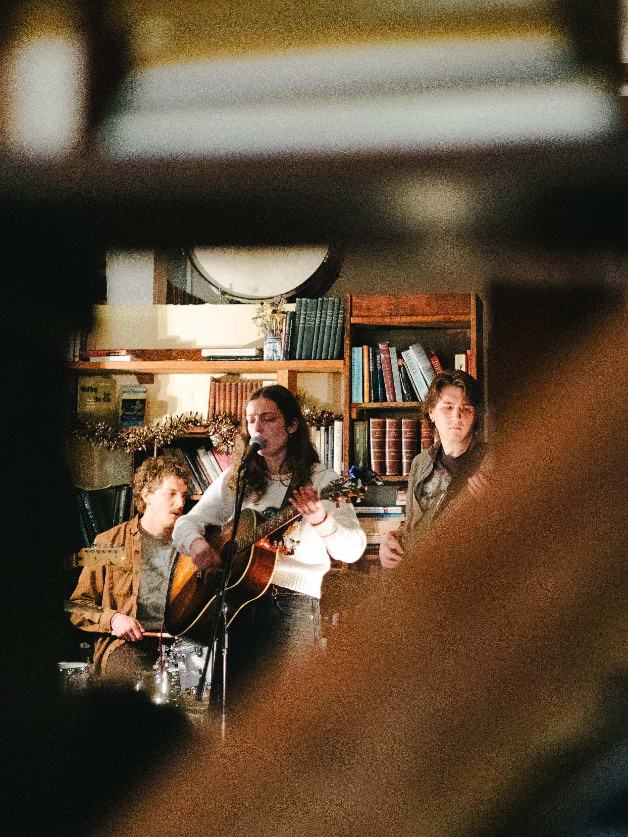 Young woman singing and playing guitar, accompanied by two young men playing other instruments, performing in a cozy room with bookshelves and holiday decorations.