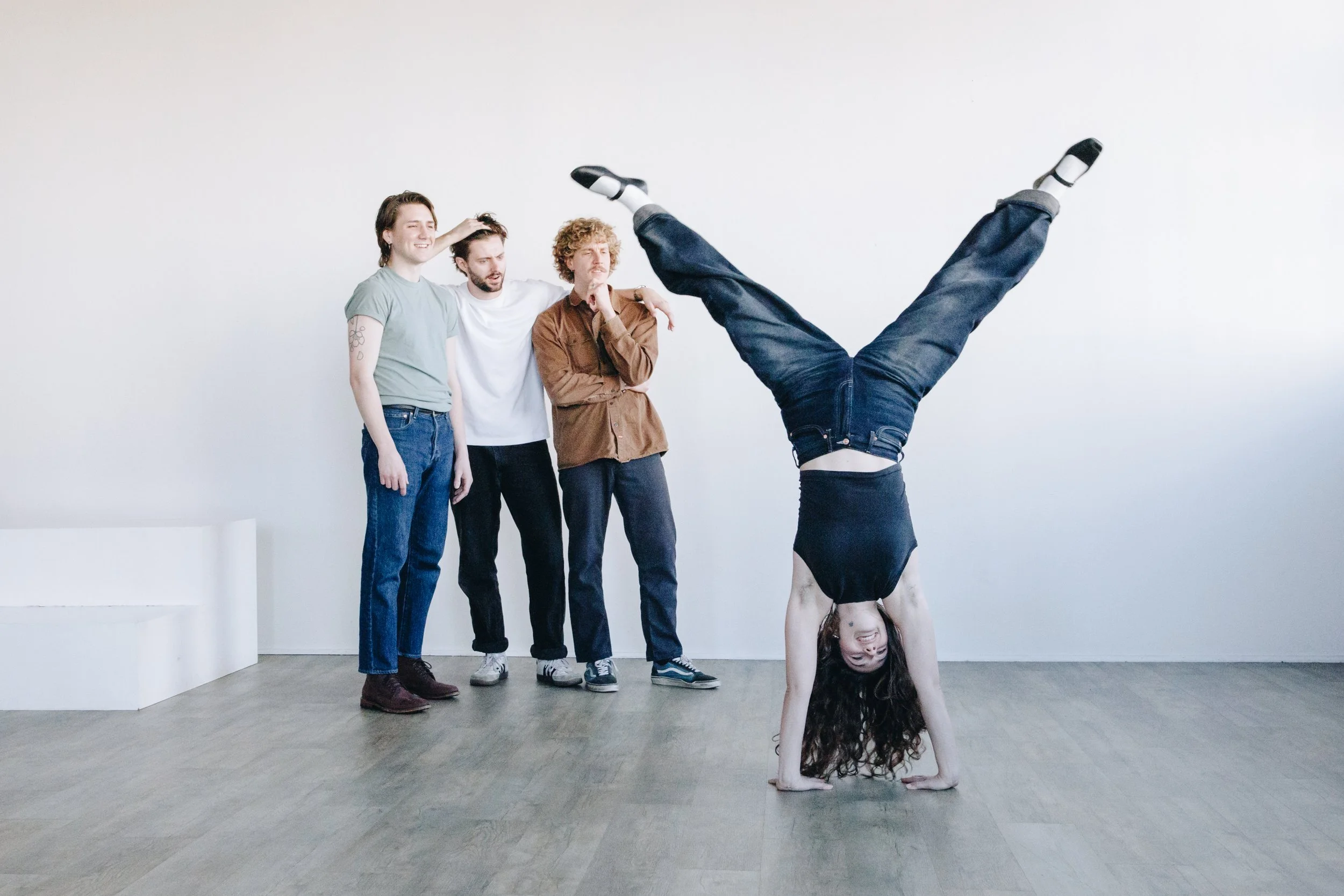 Four young adults standing against a white wall, with one person doing a handstand.