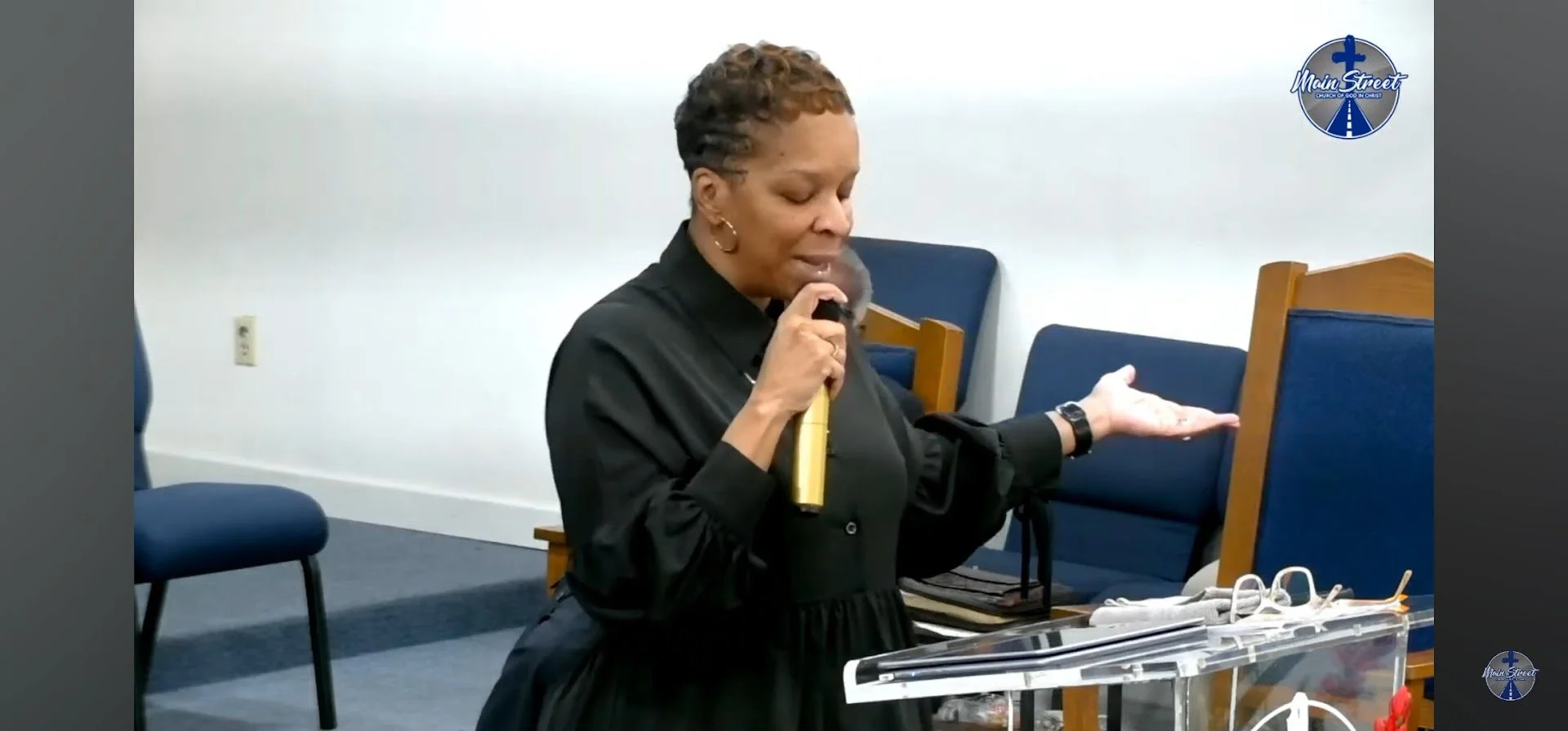 A woman with short, curly hair wearing a black dress stands at a clear acrylic podium holding a microphone in her right hand and gesturing with her left hand, in a room with blue chairs and a white wall. There is a logo with a cross and road design in the top right corner that reads 'Main Street Church of God in Christ'.