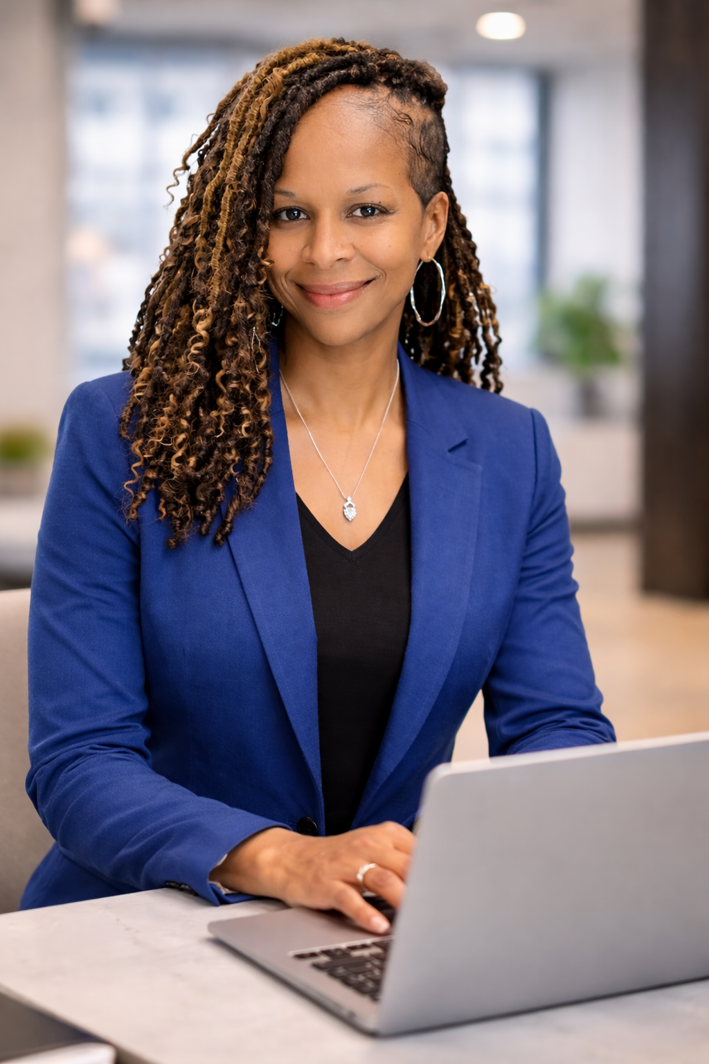 A woman in a blue blazer sitting at a desk with a laptop, smiling at the camera in an office setting.
