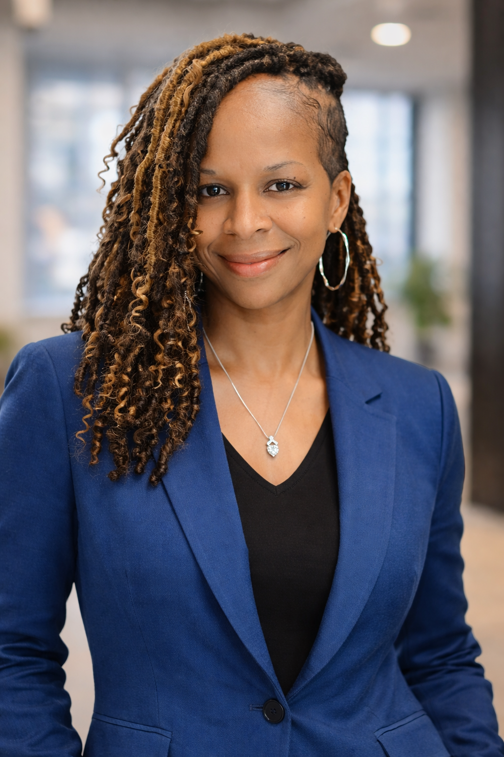A smiling woman with long curly hair wearing a blue blazer, black top, and silver jewelry in an office setting.