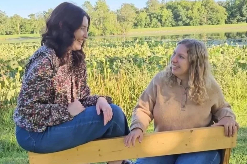 Two women smiling outdoors by a pond with lily pads, on a sunny day. Consignment boutique clothes.