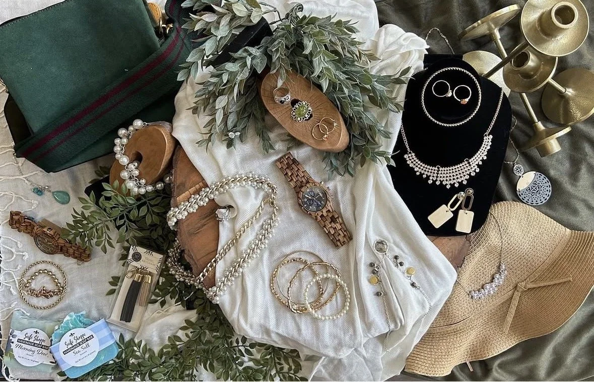 Assorted jewelry including earrings, necklaces, bracelets, rings, a watch, and pearls, displayed on a white cloth, with a green handbag, beige hat, and decorative green leaves, among other items.
