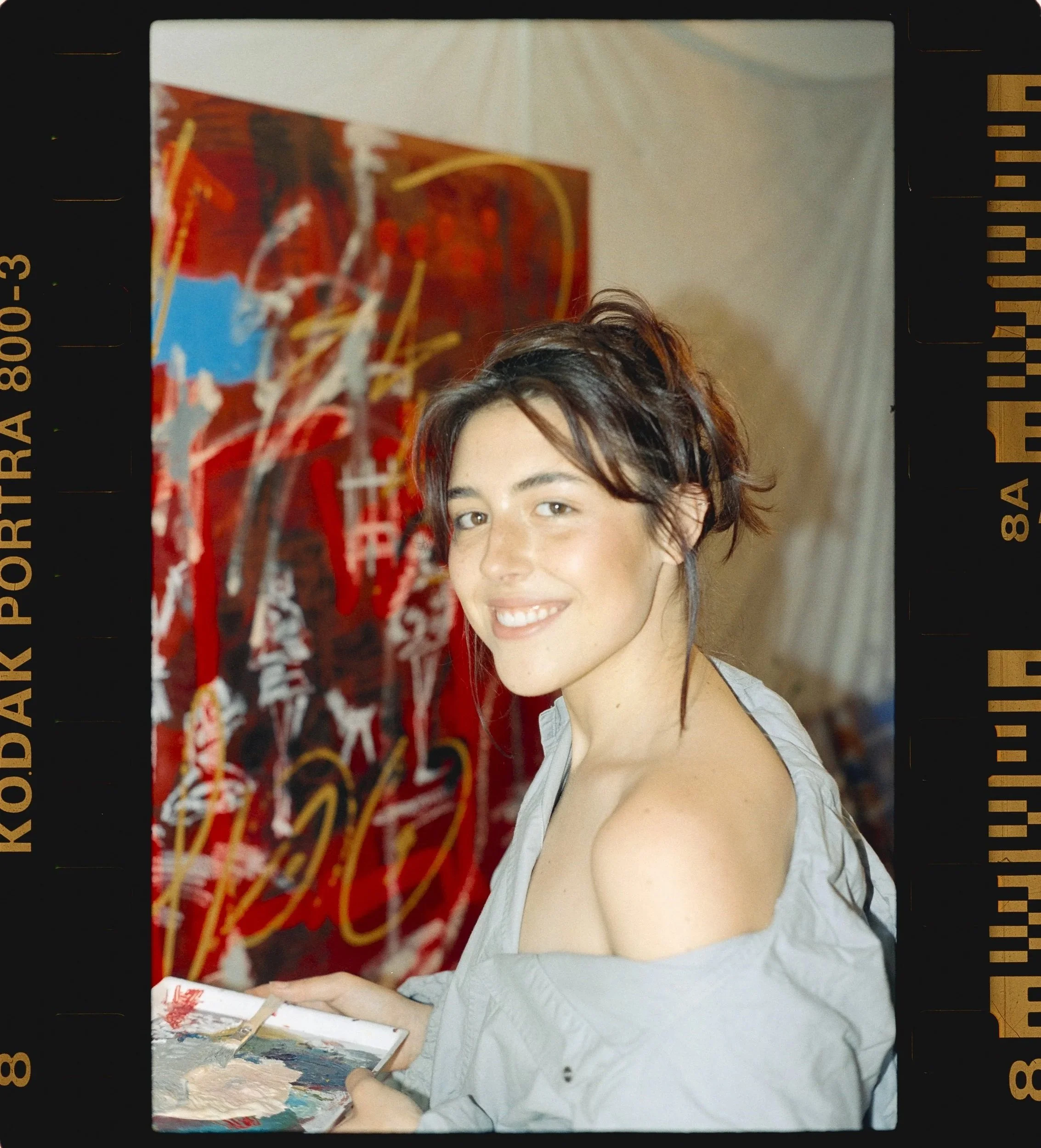 A young woman with short, dark hair smiling while holding a painter's palette and brush, standing in front of a colorful abstract painting.