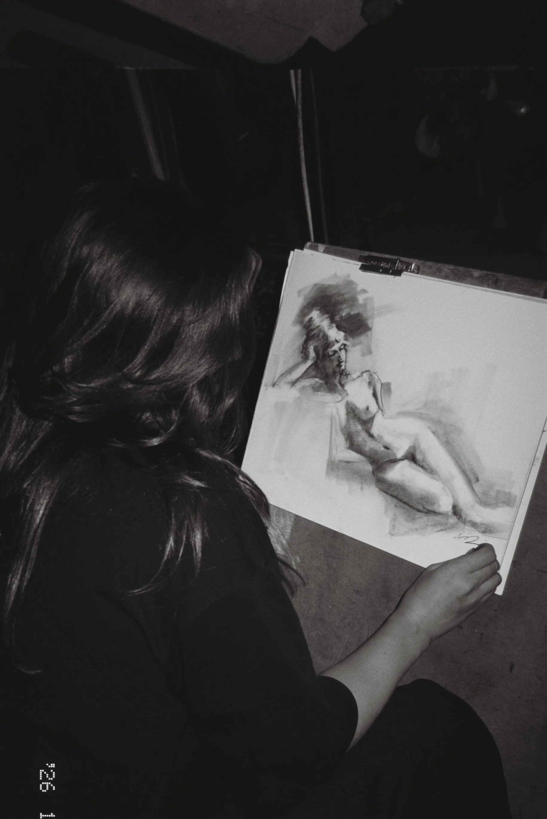 A woman with long hair is sitting and drawing a grayscale portrait of a woman on a canvas, using charcoal or pencil, in a dimly lit indoor space.
